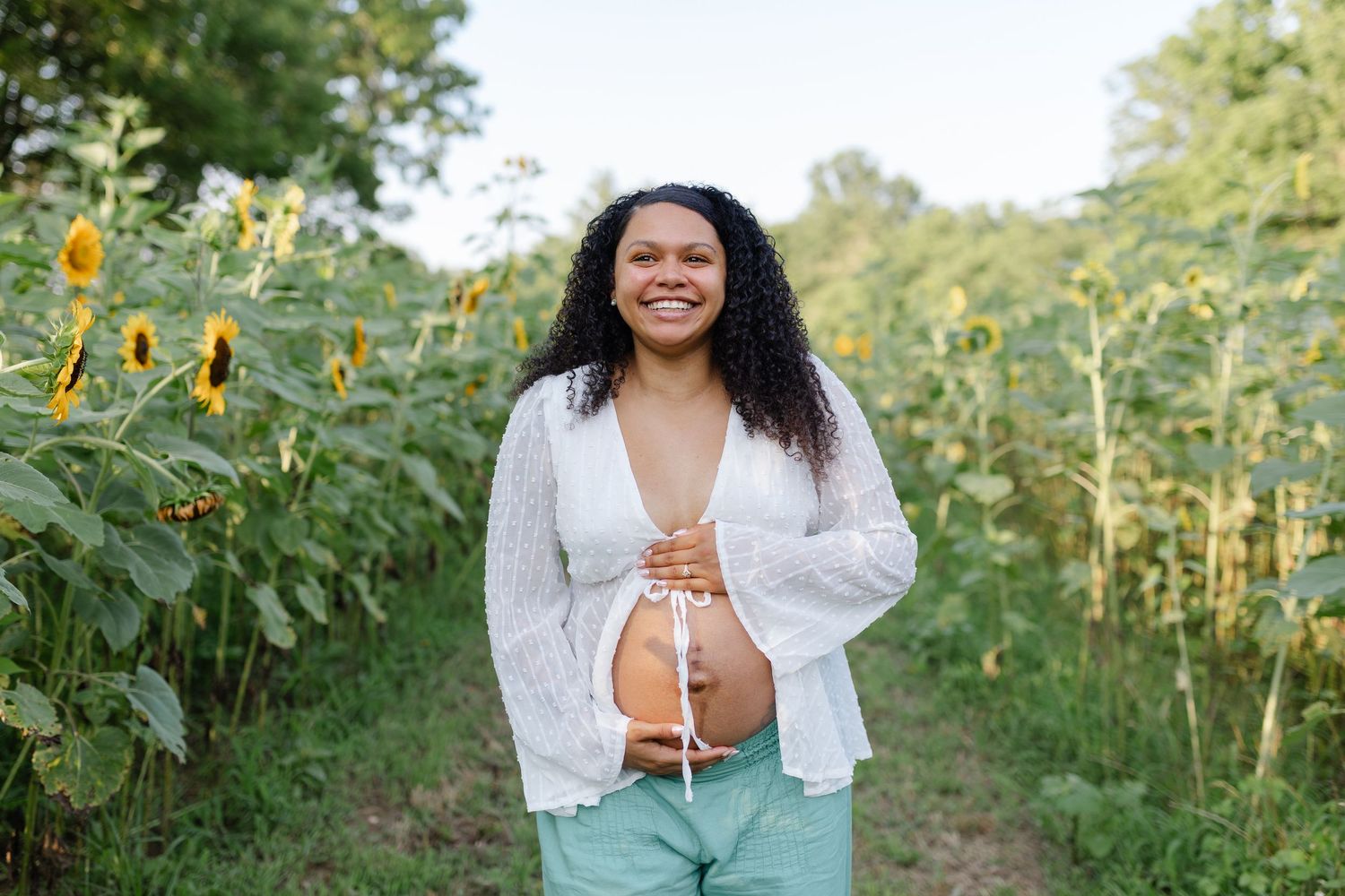 Joyful pregnant woman in white cardigan standing in sunflower field holding her belly.