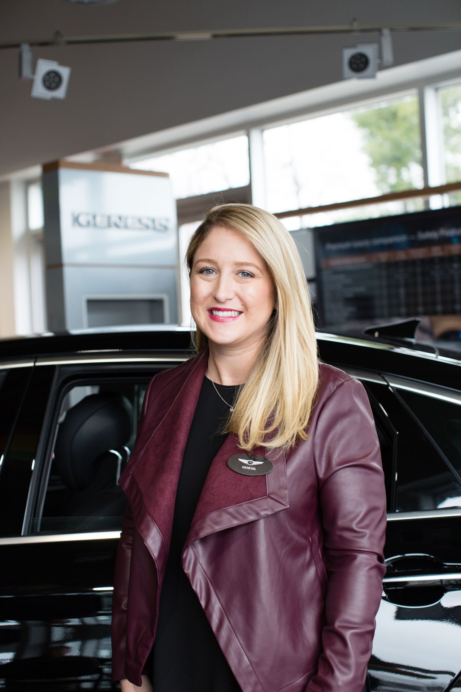 Professional in burgundy leather jacket standing near luxury vehicles in showroom.