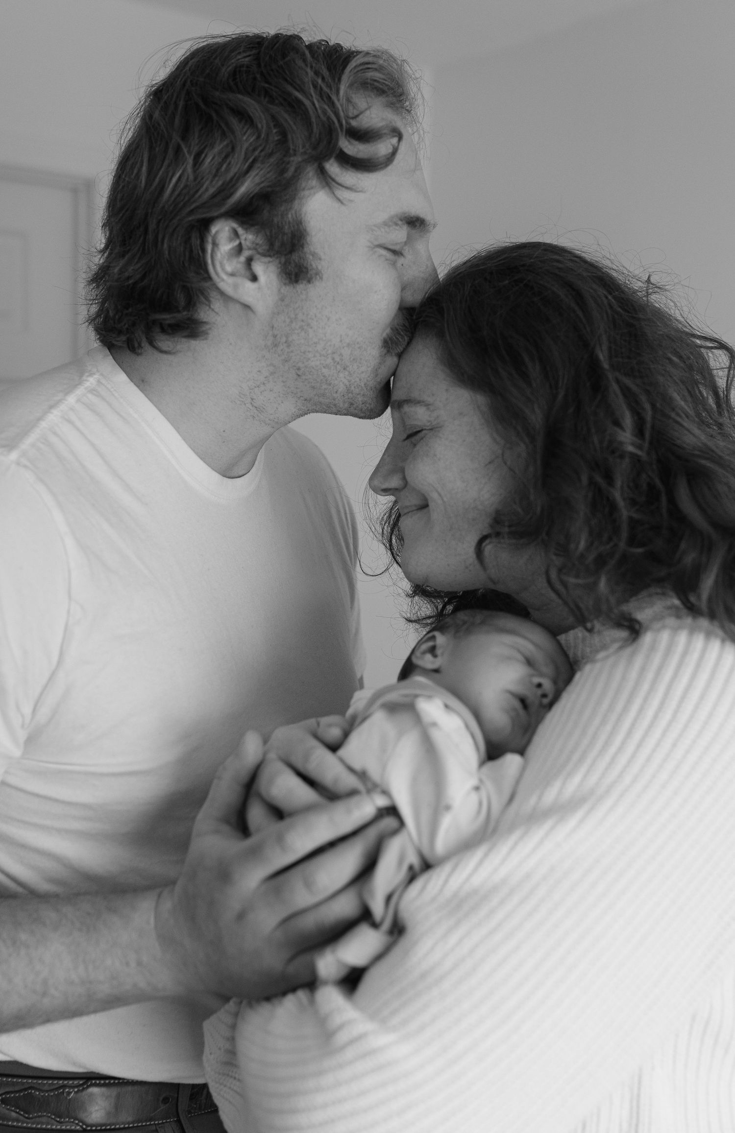 Black and white photo of new parents sharing a tender kiss over their sleeping newborn baby.