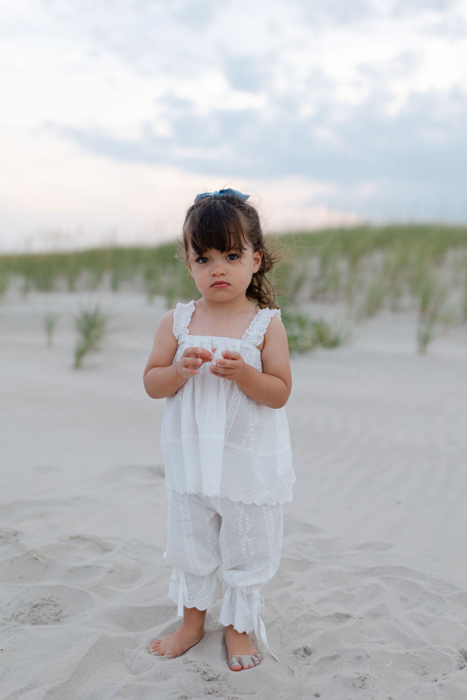 Adorable toddler girl in white dress and pants standing barefoot on sandy beach.
