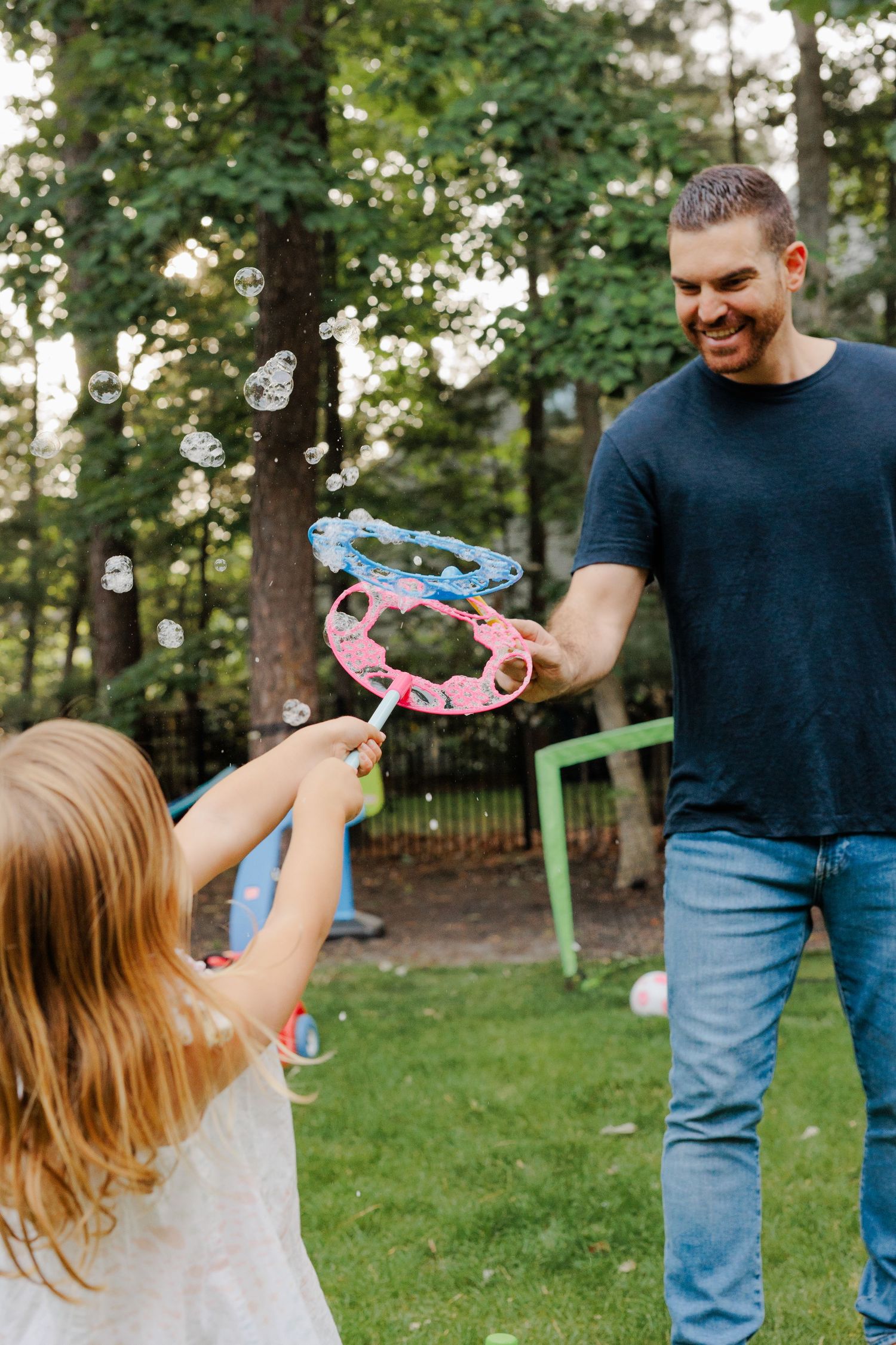 Smiling father playing with bubble wands with young blonde daughter in a green backyard.