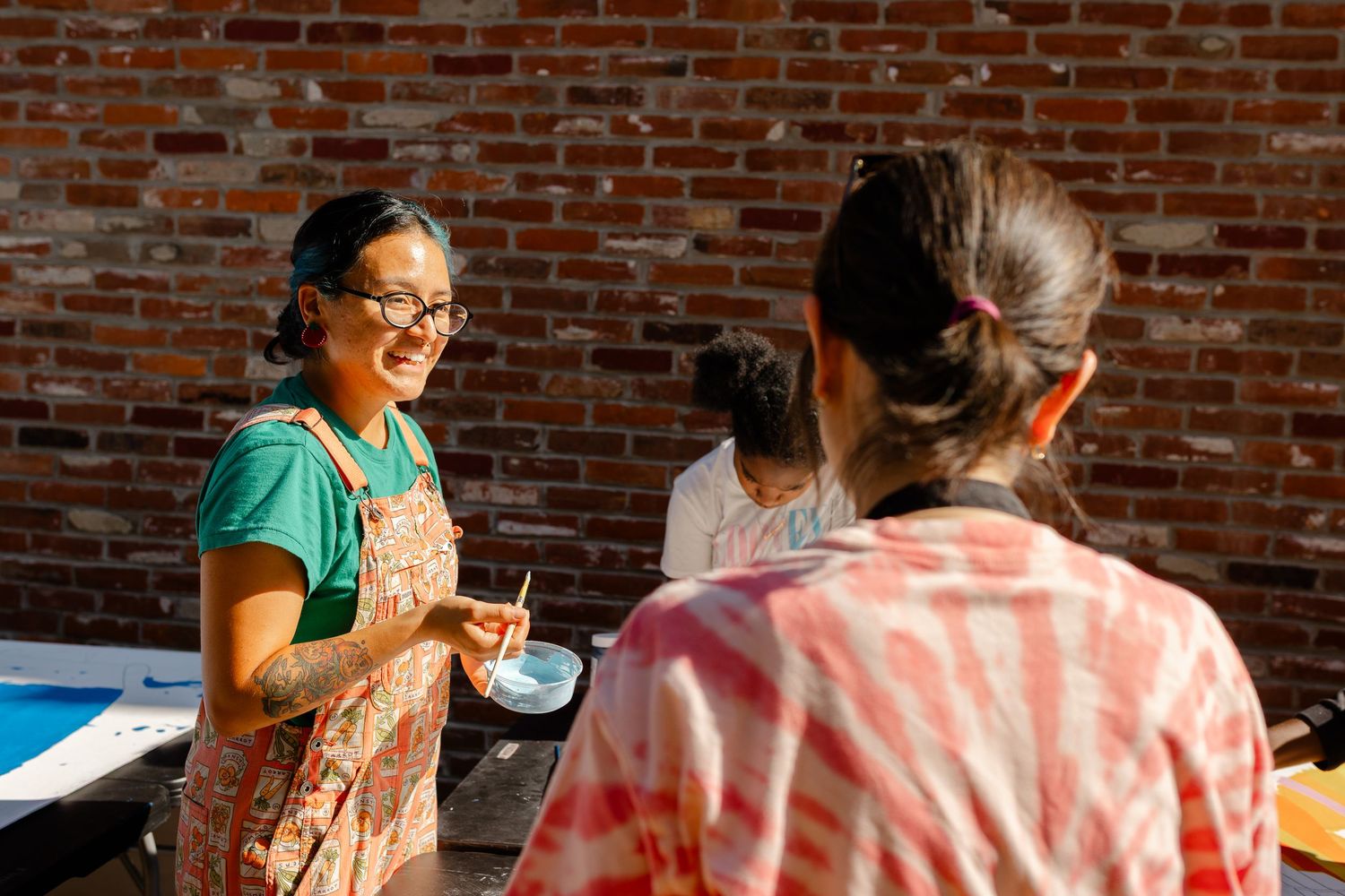 Two women in colorful aprons talk and smile outdoors near a brick wall during an art workshop.