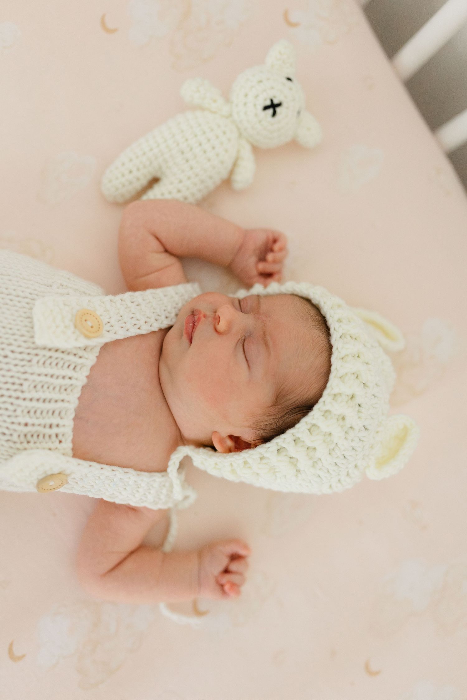 Sleeping newborn baby in white crochet bear outfit beside a knitted bunny toy on a pink crib sheet.