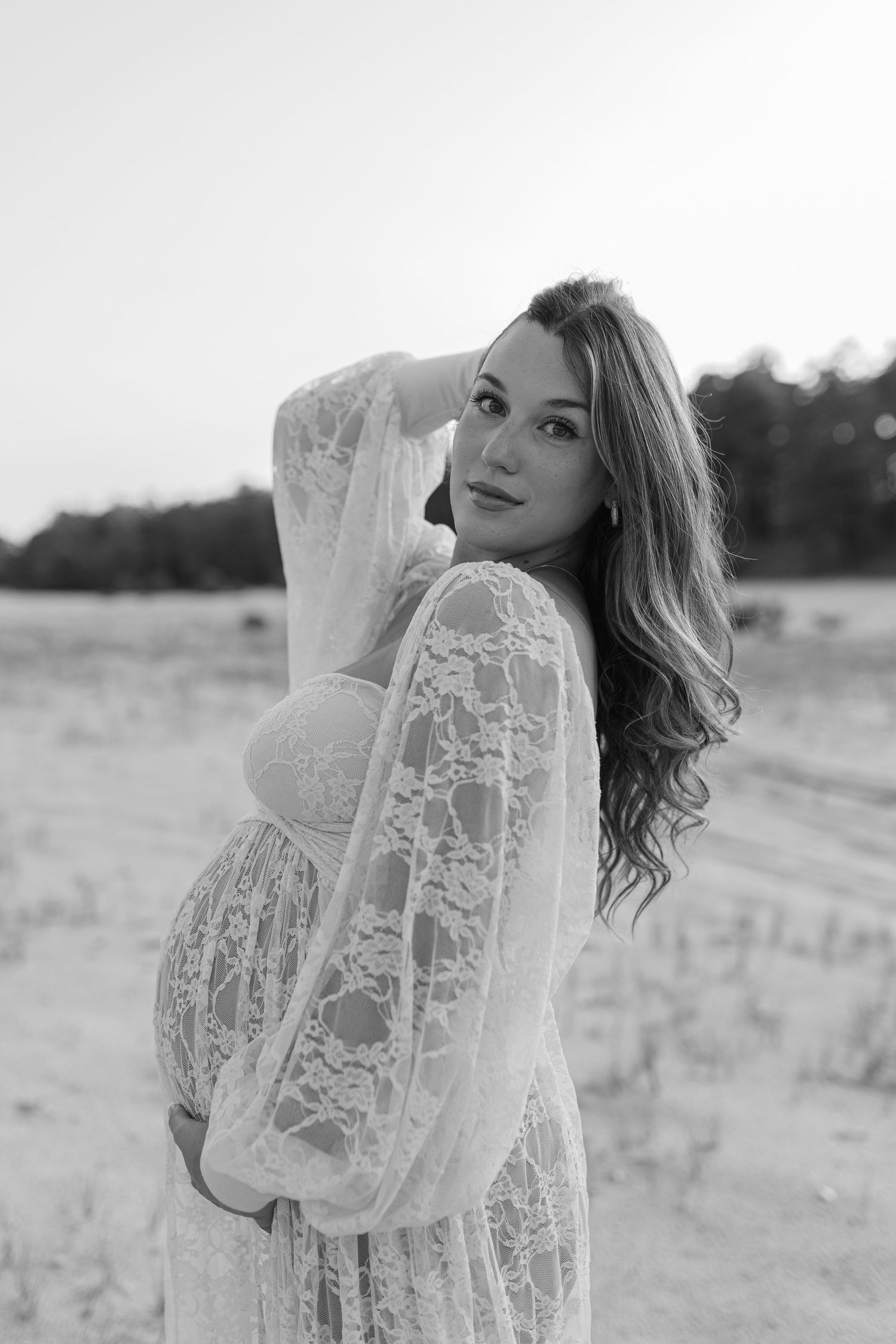 Pregnant woman in white lace maternity gown poses gracefully on sandy beach in black and white photo.