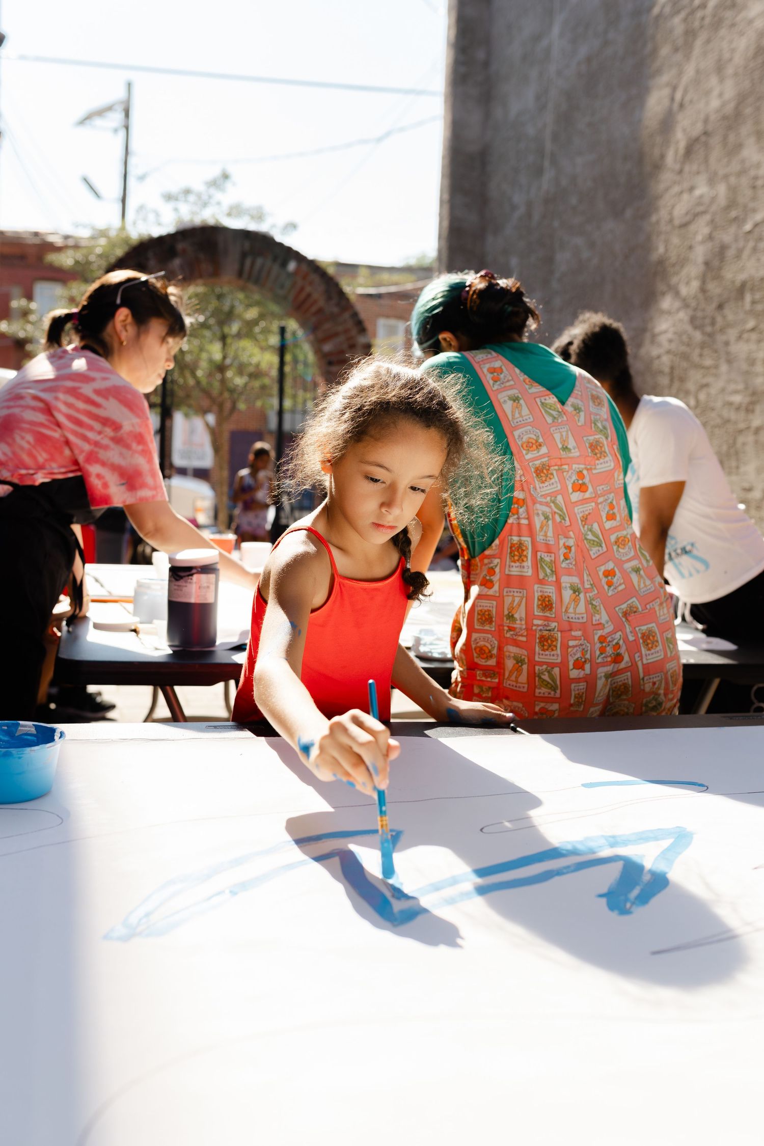 Young girl in red top painting blue artwork on white paper at outdoor community art event.