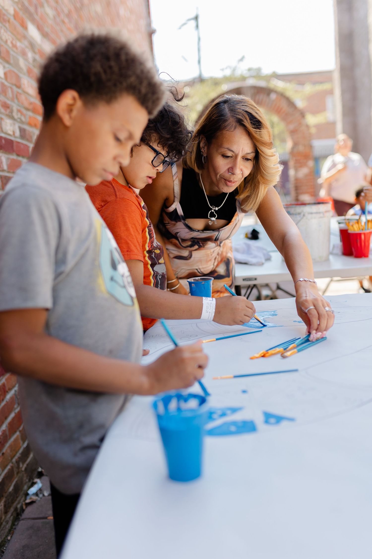 Woman and two children painting with blue paint at an outdoor art activity table near a brick wall.