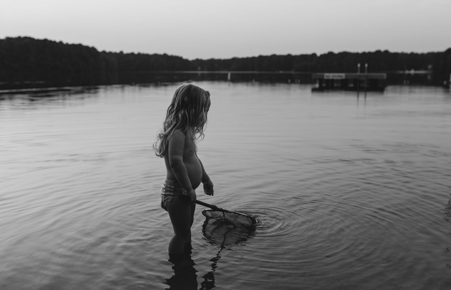 Young girl wading in a lake at dusk holding a fishing net in a black and white photo.