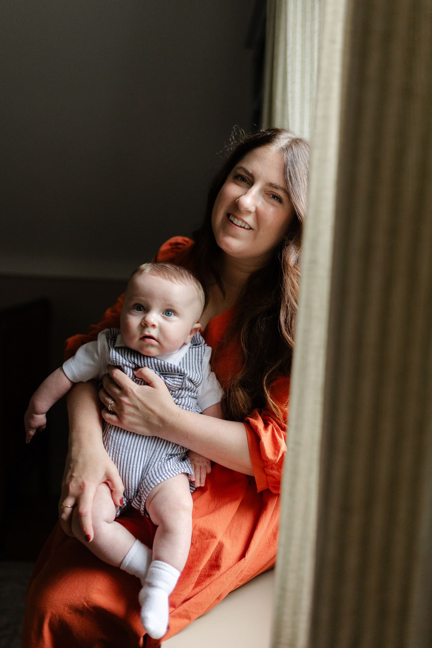 Smiling mother in orange dress holding her baby boy near a window in soft natural light.