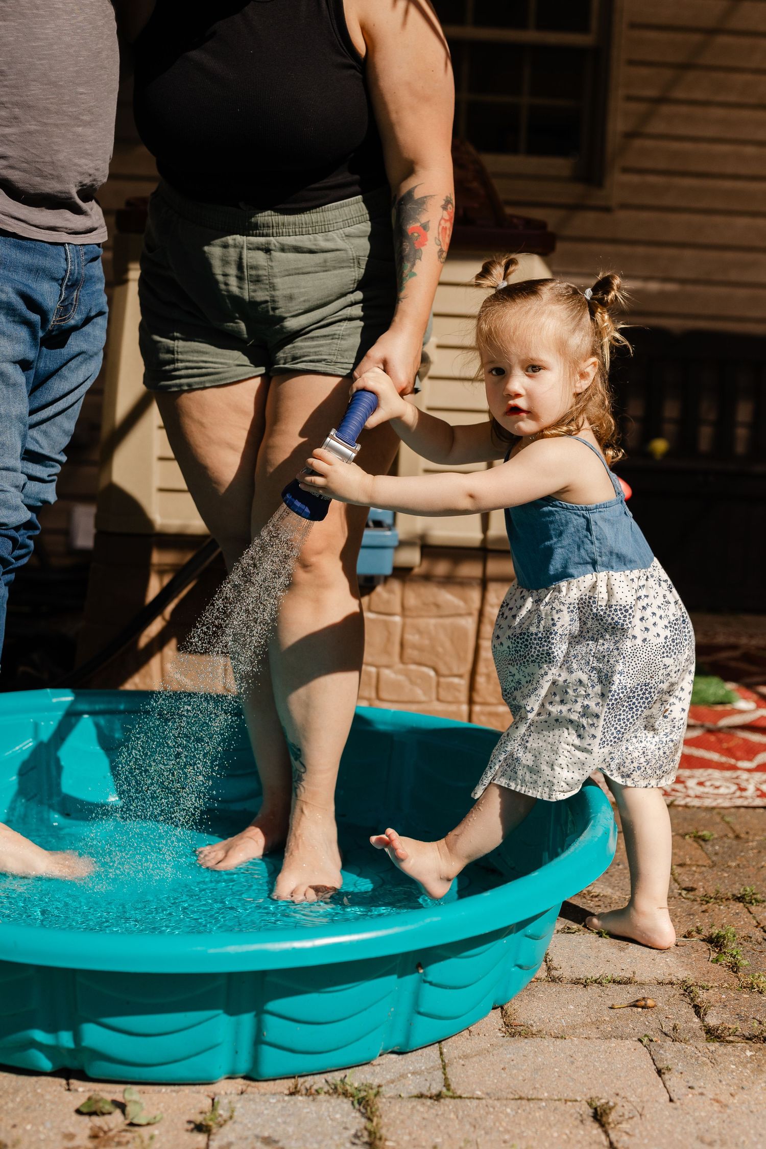 Toddler girl standing in a turquoise kiddie pool with adults helping her play with water on a sunny day.