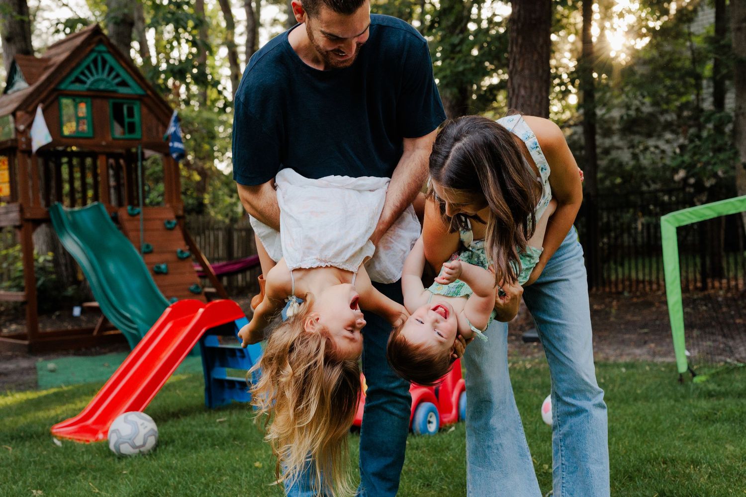 Parents playfully dipping two young children upside down together in a green backyard.