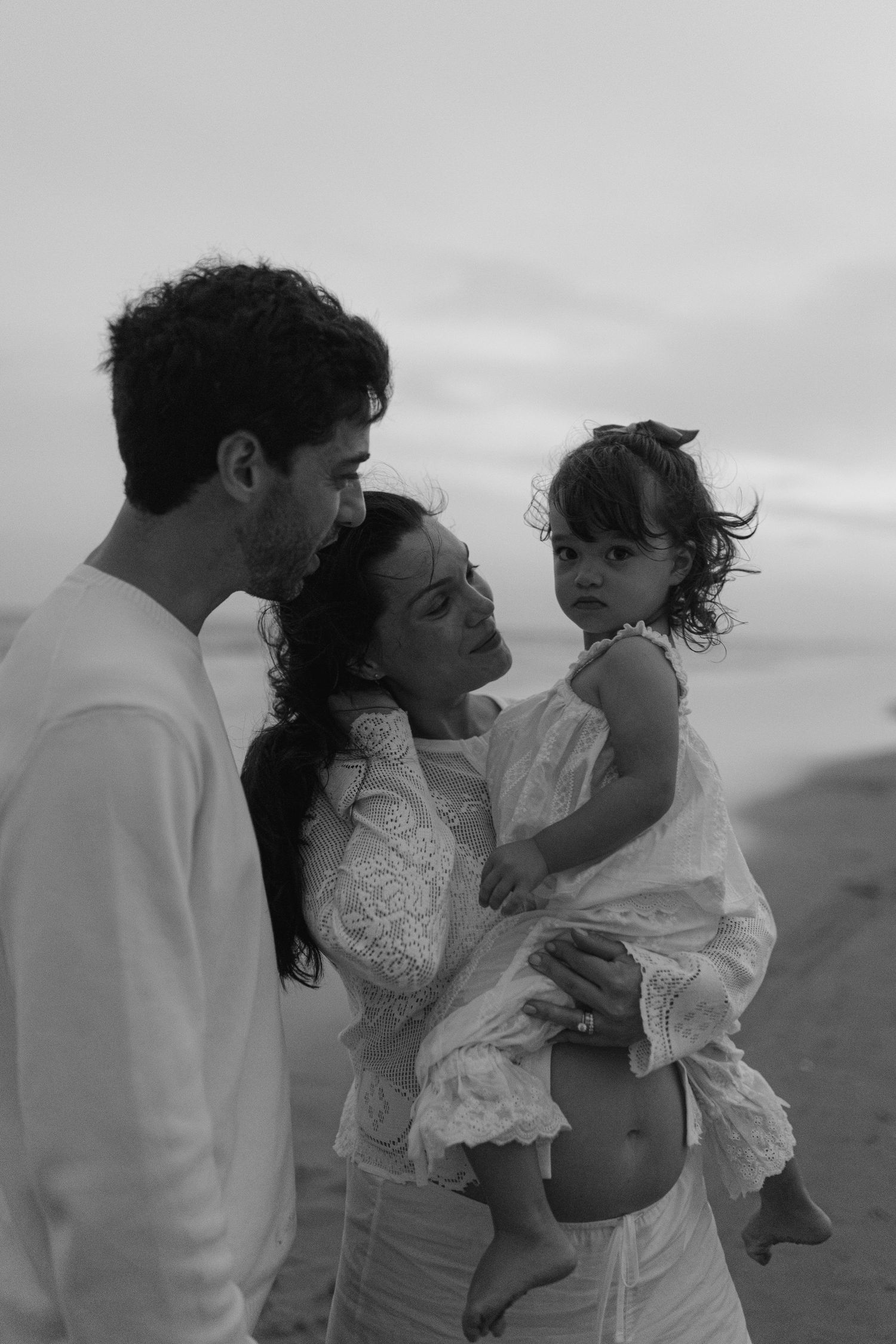 Black and white photo of a couple with their toddler daughter on a beach at dusk, all dressed in white.