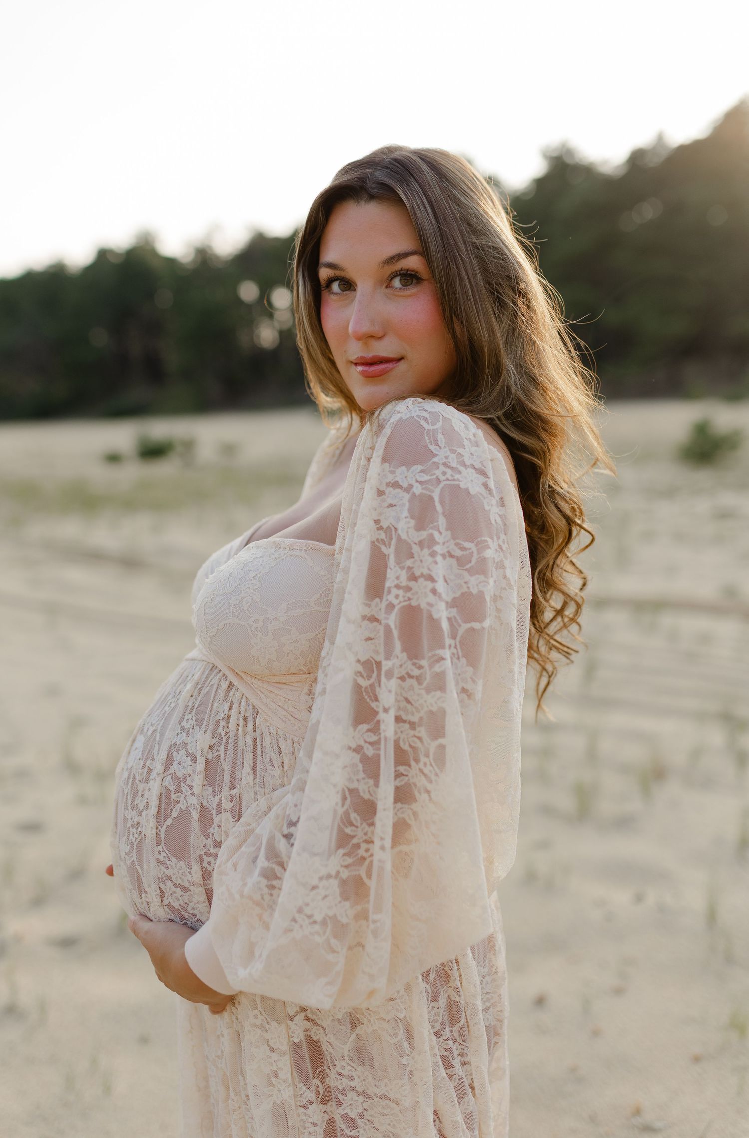 Pregnant woman in white lace dress posing in sandy field at golden hour.