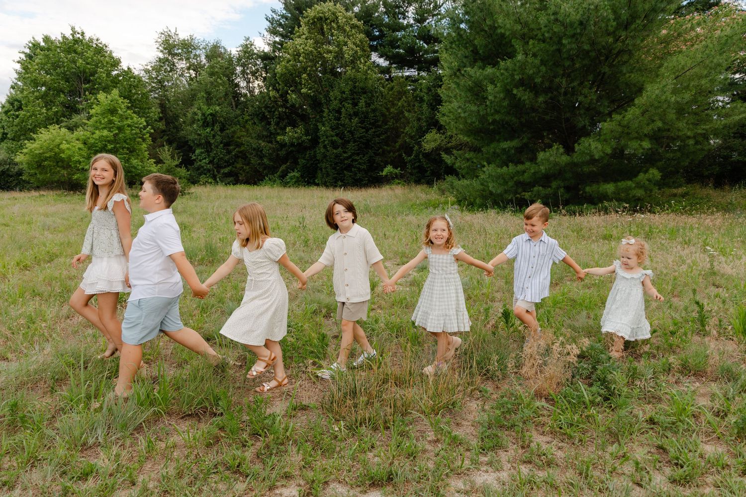 Seven children in white outfits hold hands walking through a green meadow.