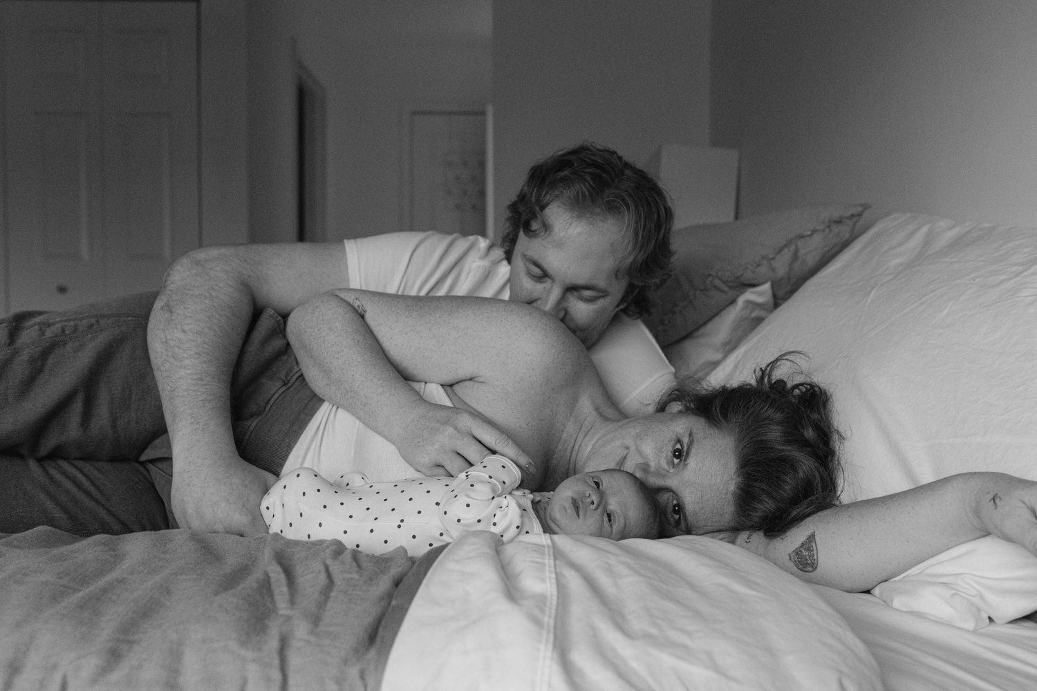 Black and white intimate photo of couple cuddling with newborn baby in bed.