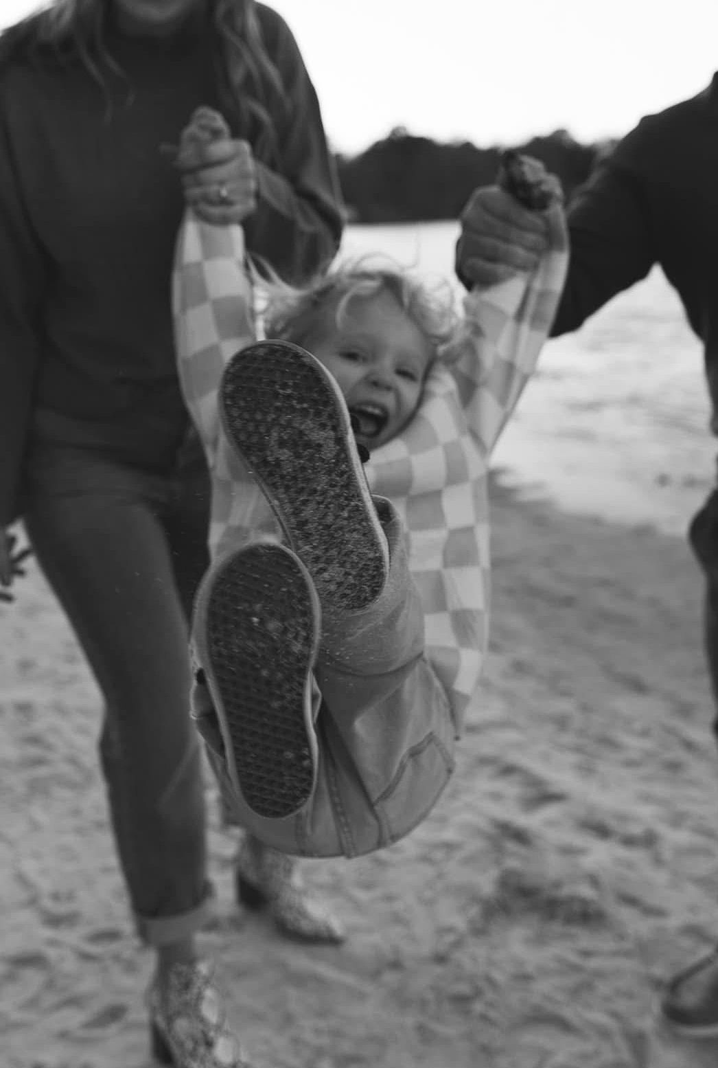 Black and white photo of parents swinging their laughing toddler on a sandy beach.