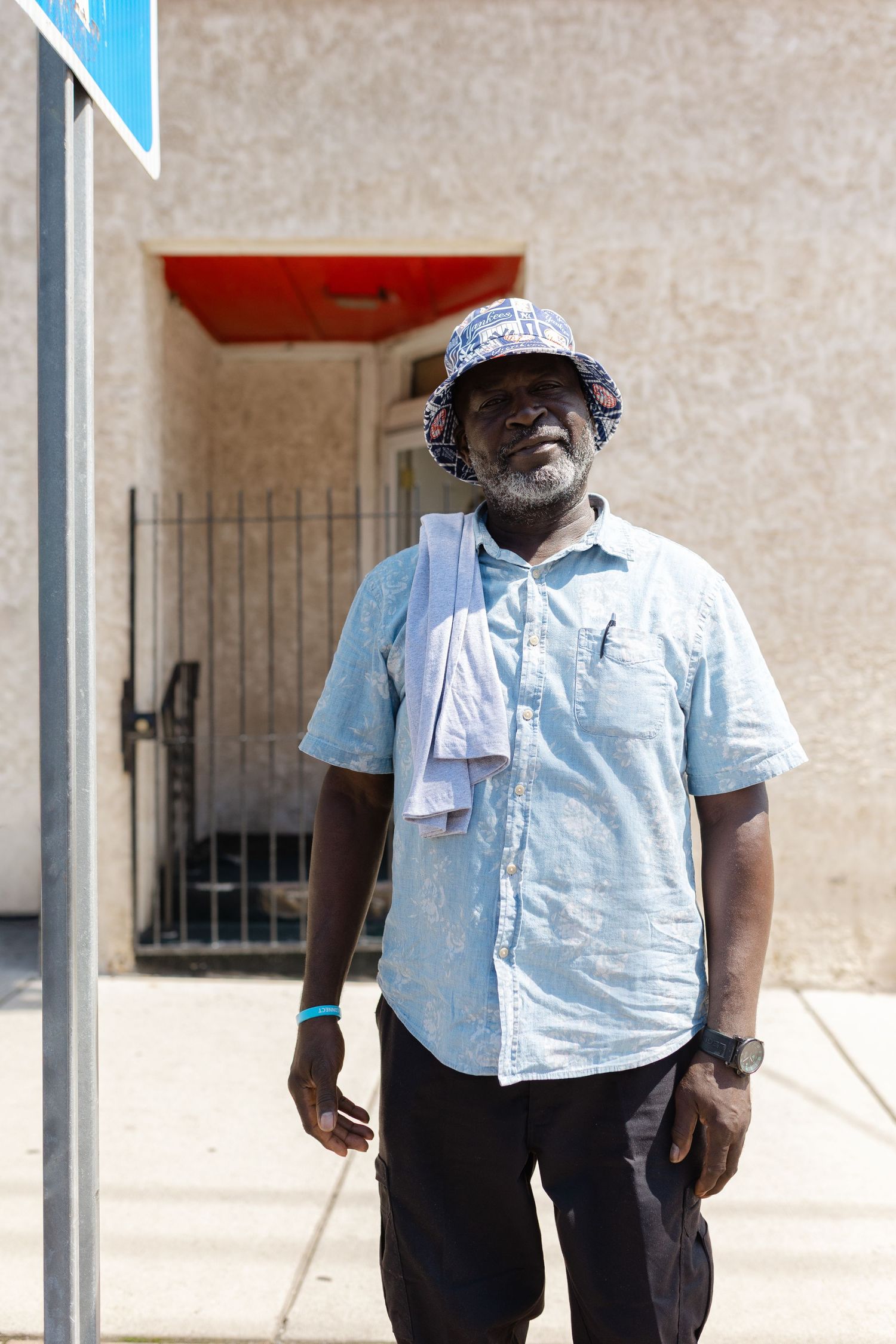 Smiling Black man in light blue shirt and bucket hat standing outdoors on a sunny day near a building.