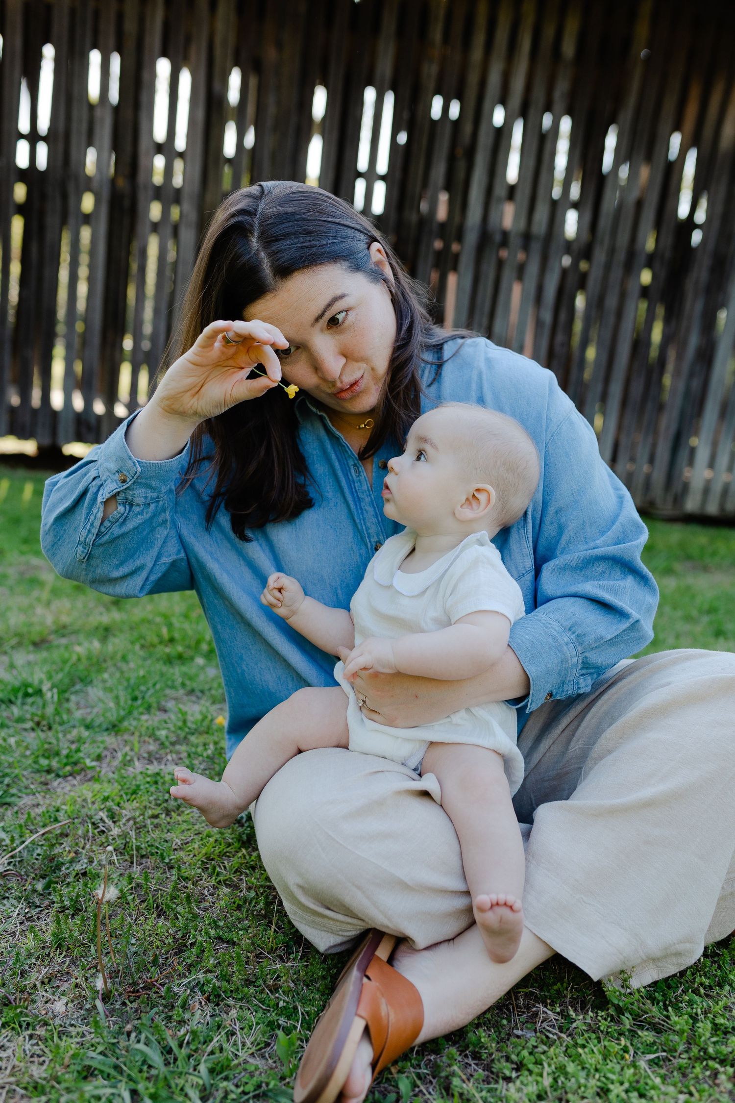 Mother sitting on lawn holding baby while playfully placing small flower near her eye.