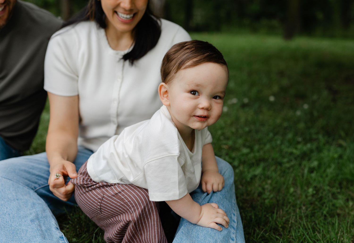 Adorable toddler in white shirt crawling on grass with mother smiling in background.