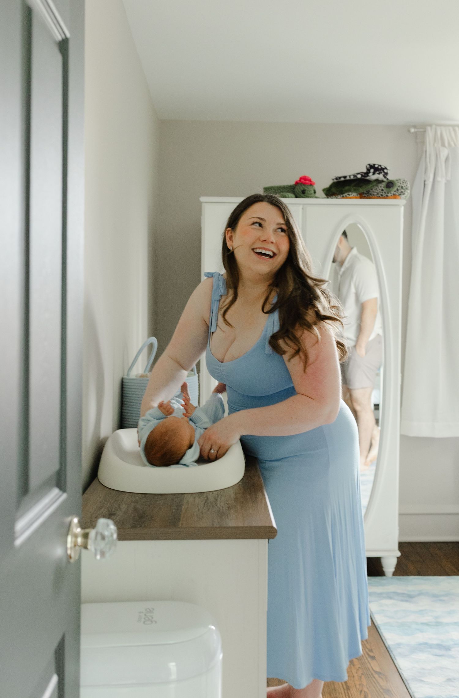 Happy new mother laughing while changing newborn baby on dresser in bright nursery room.