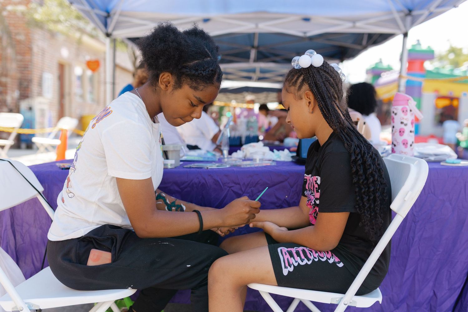 Two girls sit together at an outdoor event table, smiling and sharing a phone under a purple canopy.