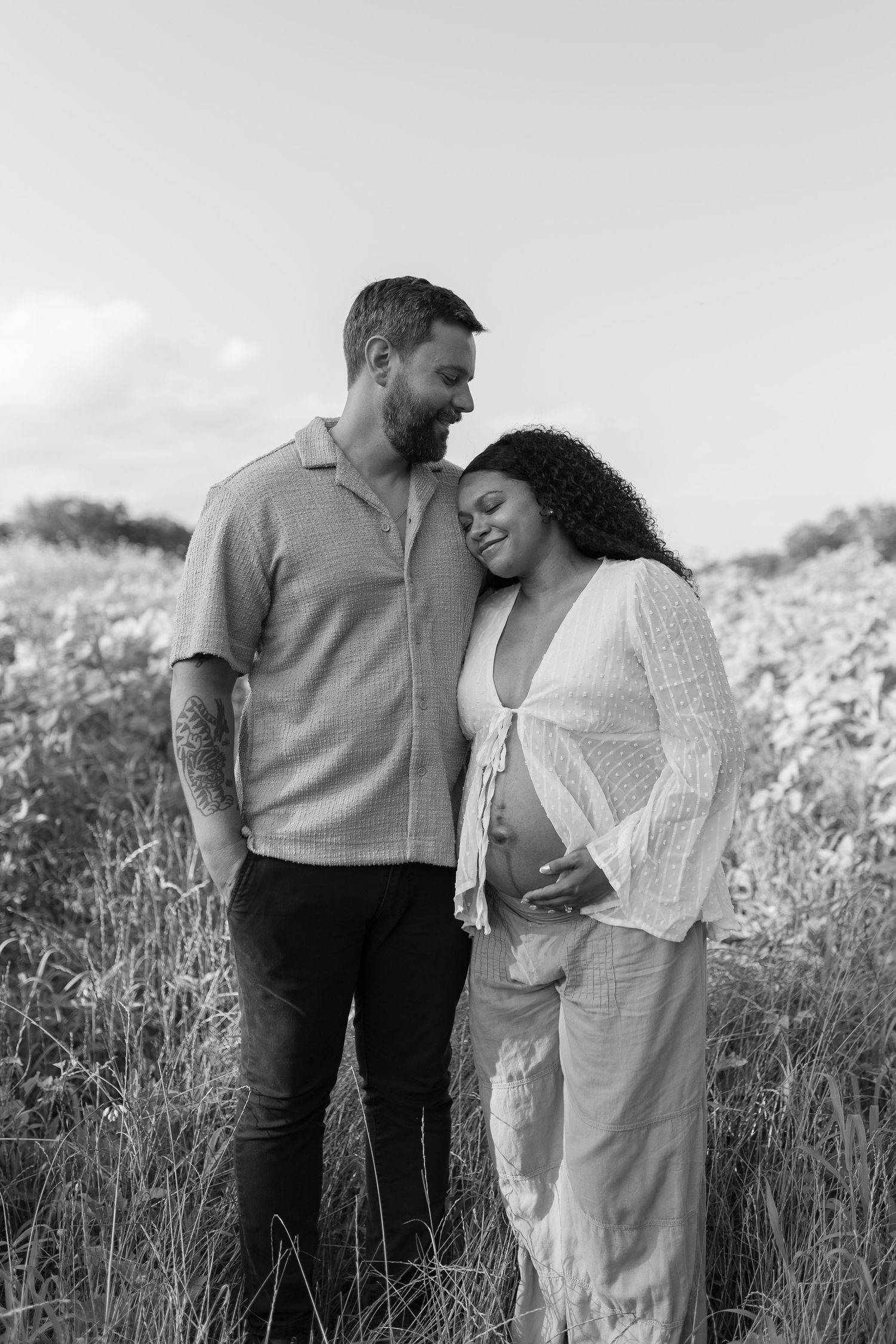 Black and white maternity photo of a couple standing together in a field, woman leaning on partner's shoulder.