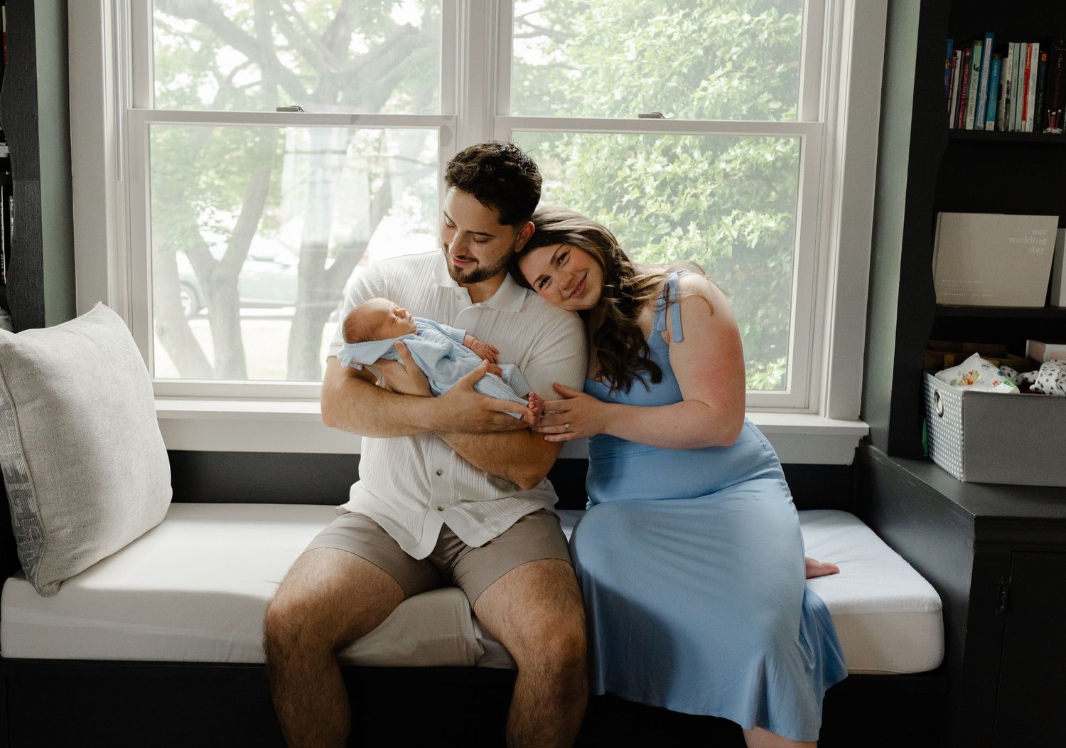 New parents sitting by window holding sleeping newborn baby, leaning heads together lovingly.