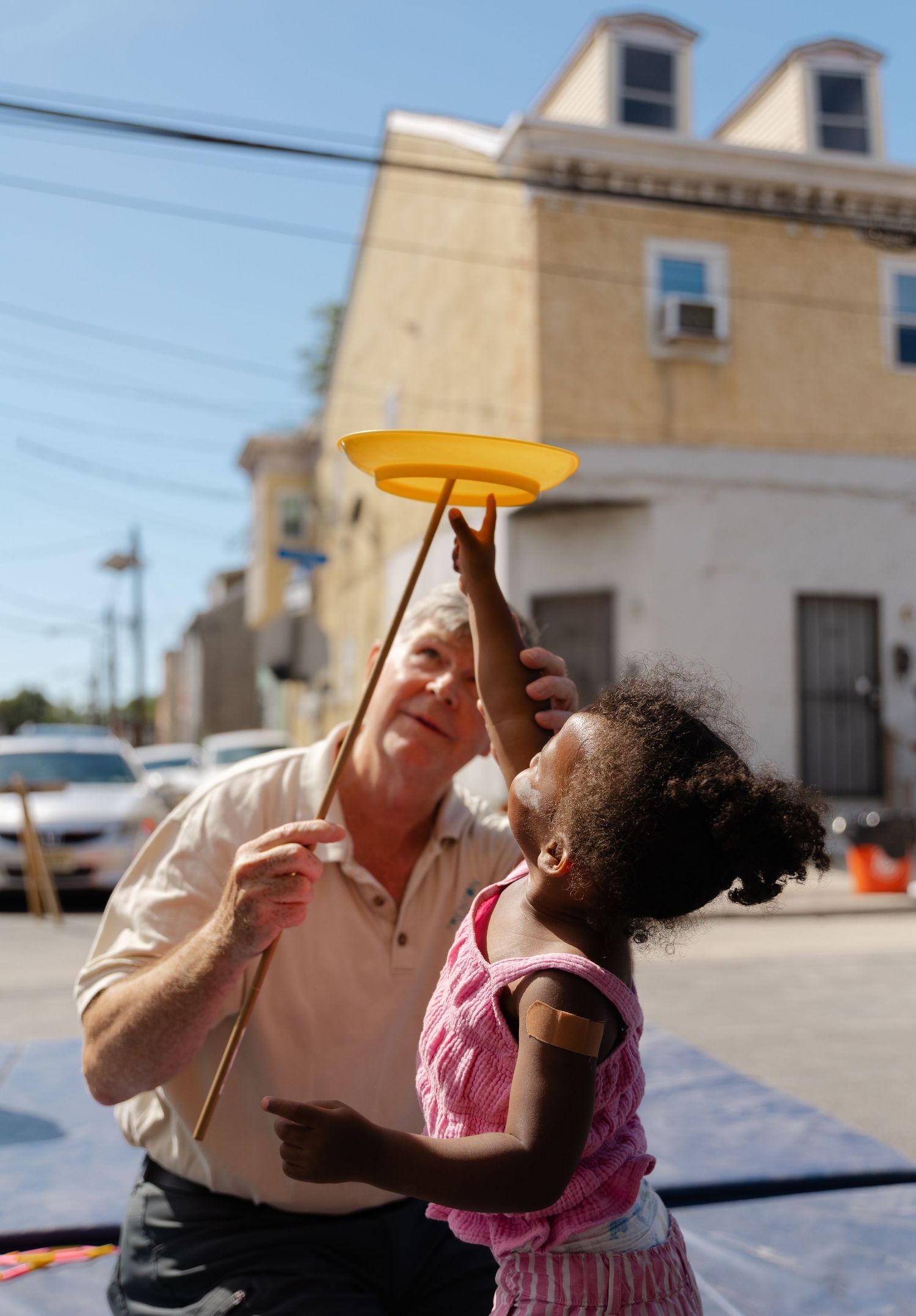 An elderly man teaches a young girl to spin a yellow plate on a stick outdoors on a sunny day.