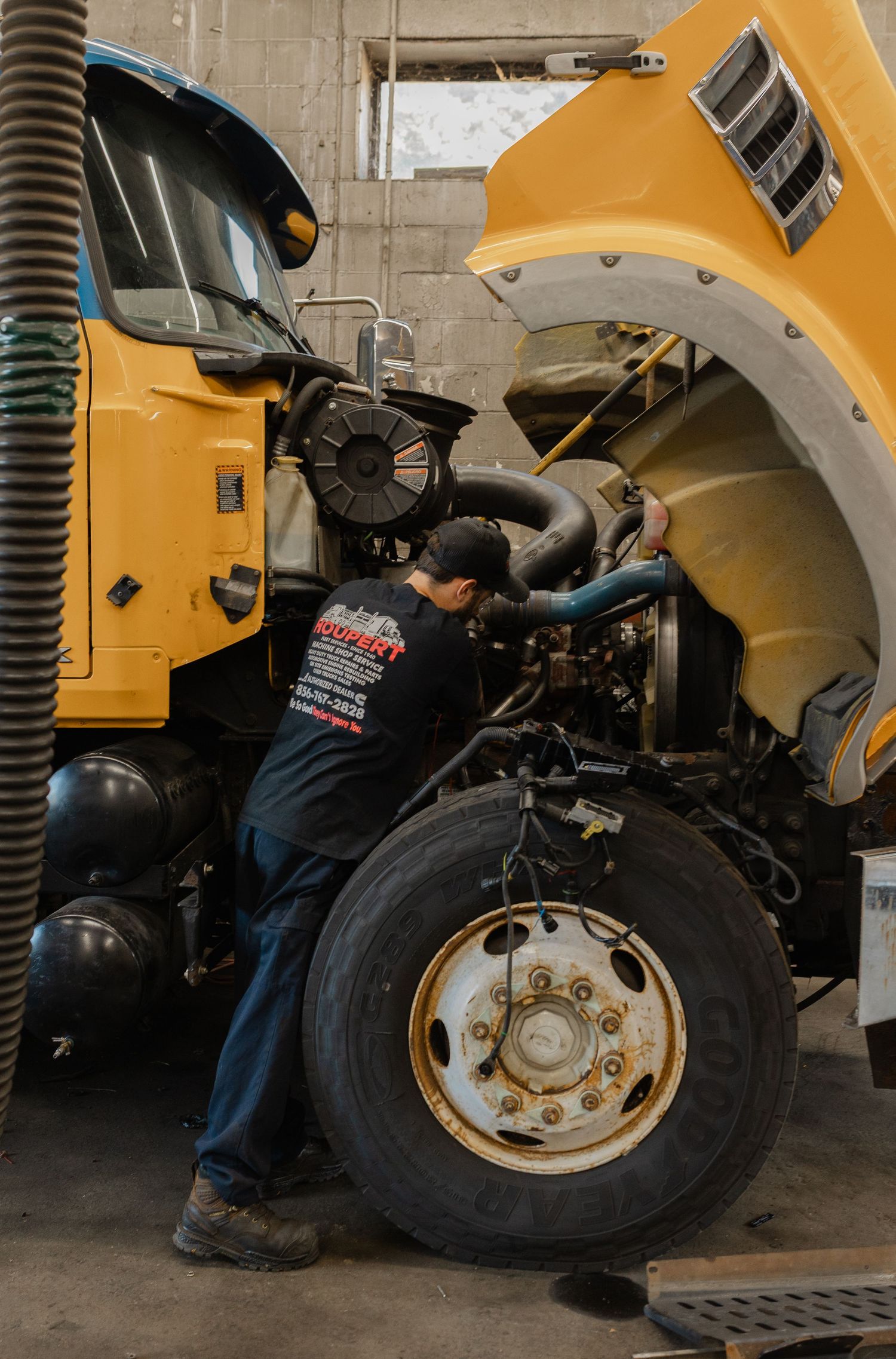 A mechanic in a black uniform repairs the engine of a large yellow commercial truck in an auto repair shop.