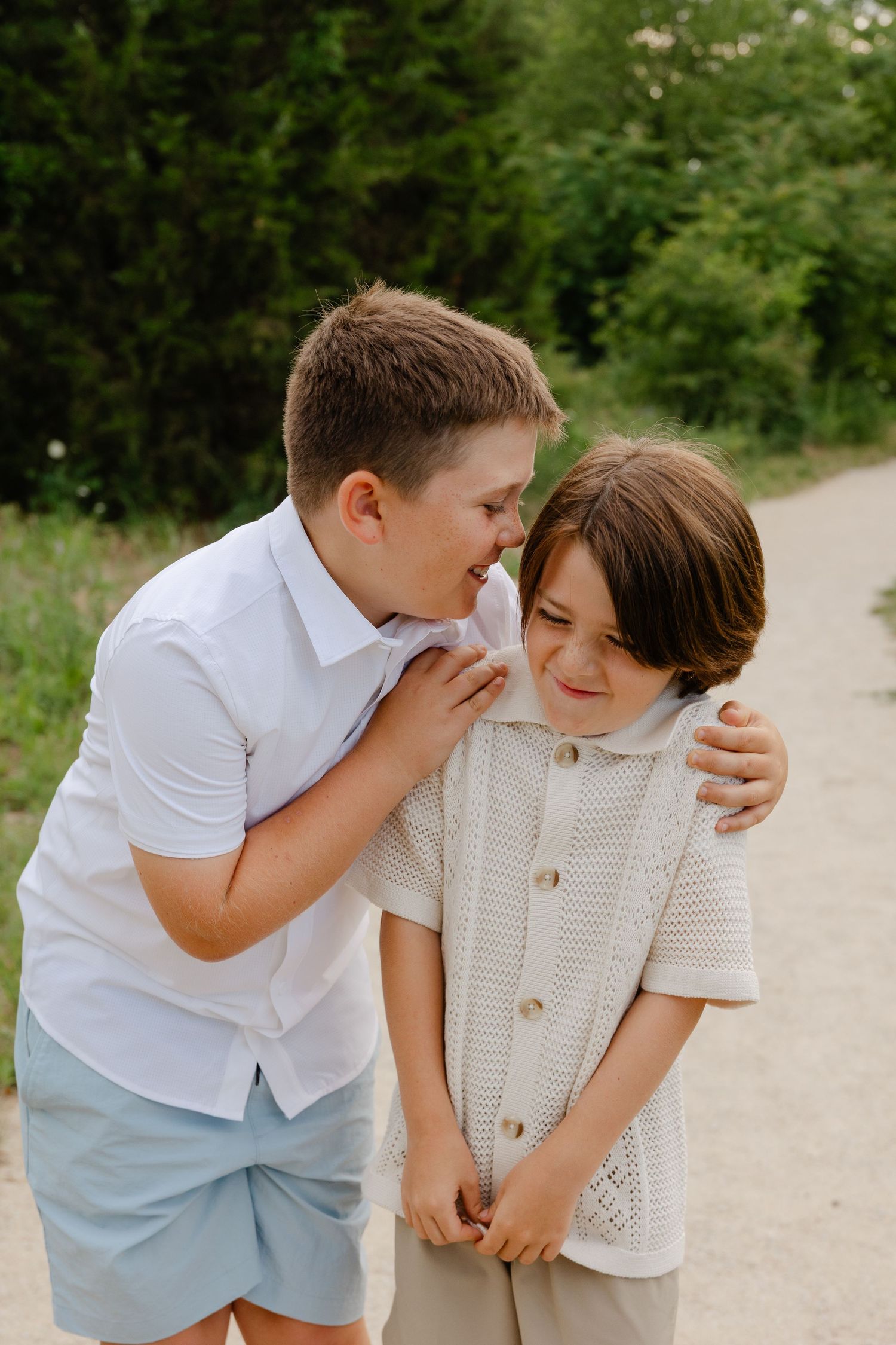 Two brothers share a laugh as older boy whispers to younger on a garden path.