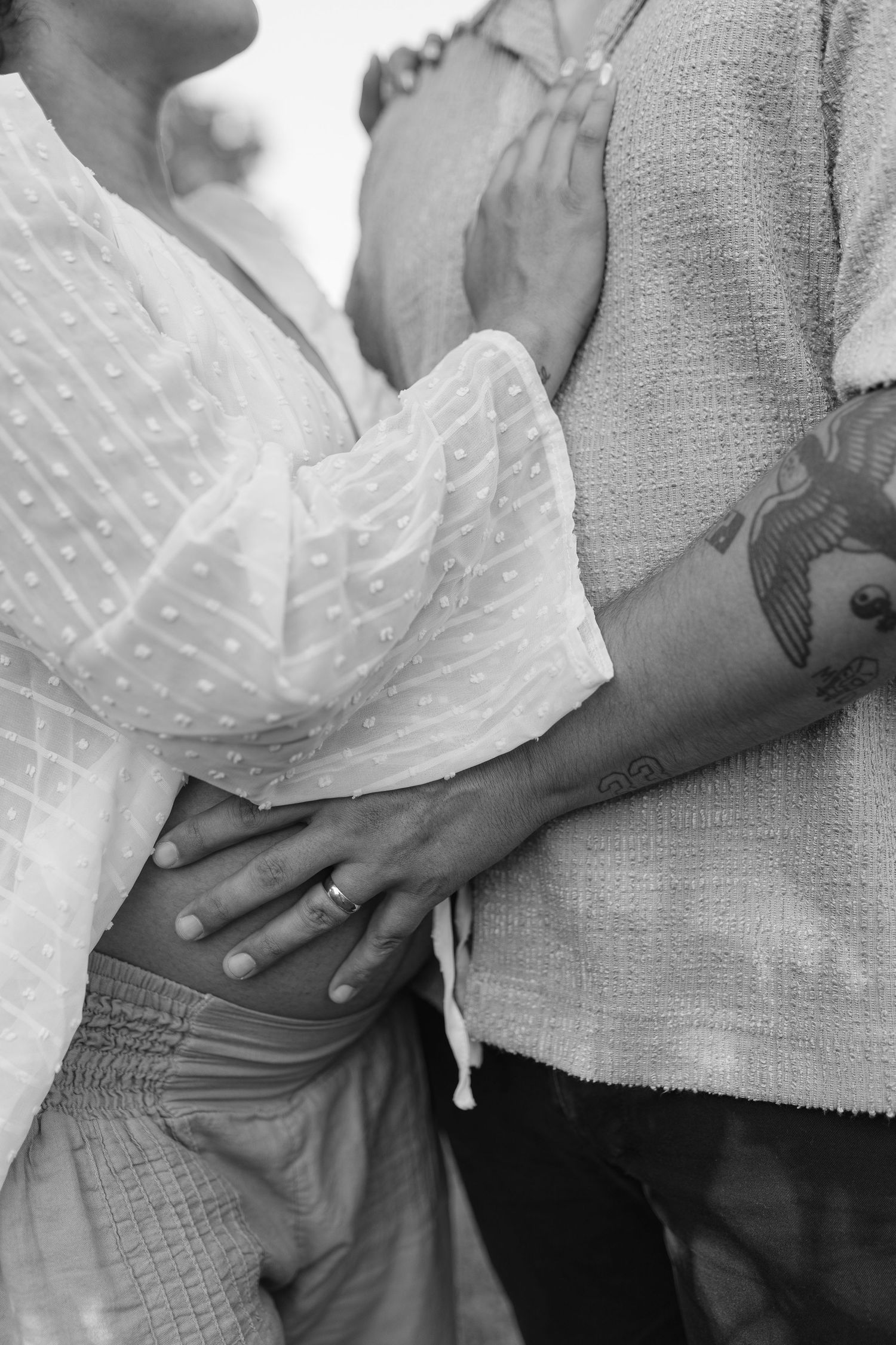 Black and white photo of a pregnant couple embracing, hands resting on baby bump, tattoo visible on partner's arm.
