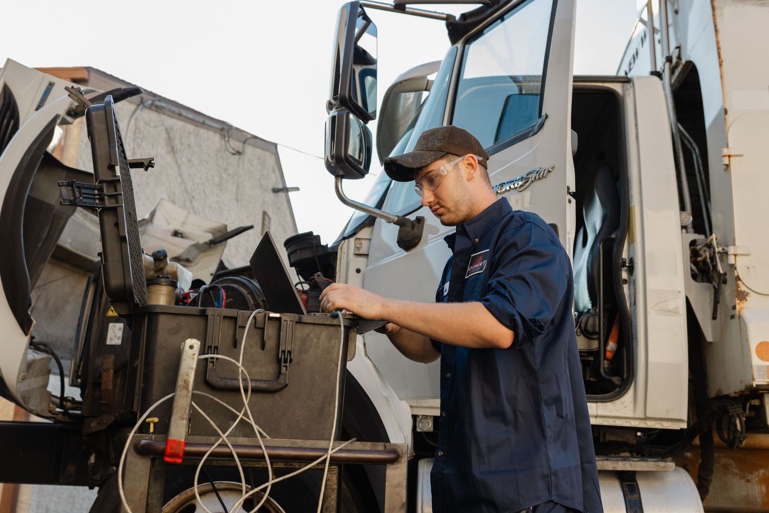 Technician in blue uniform inspecting heavy truck equipment outdoors, performing diesel vehicle maintenance and diagnostics.