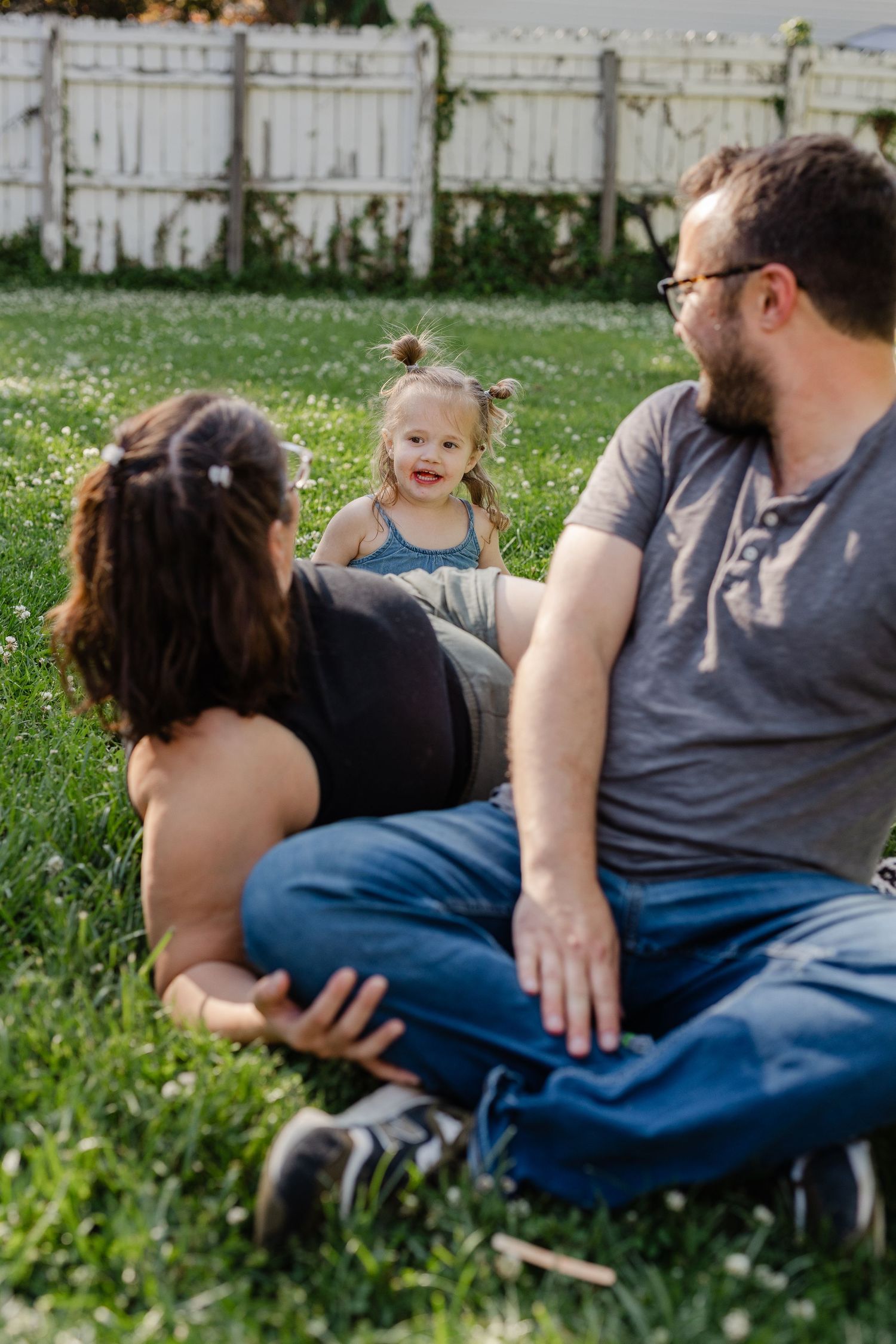 Young toddler girl smiling at her parents while sitting together on a clover-filled lawn in backyard.