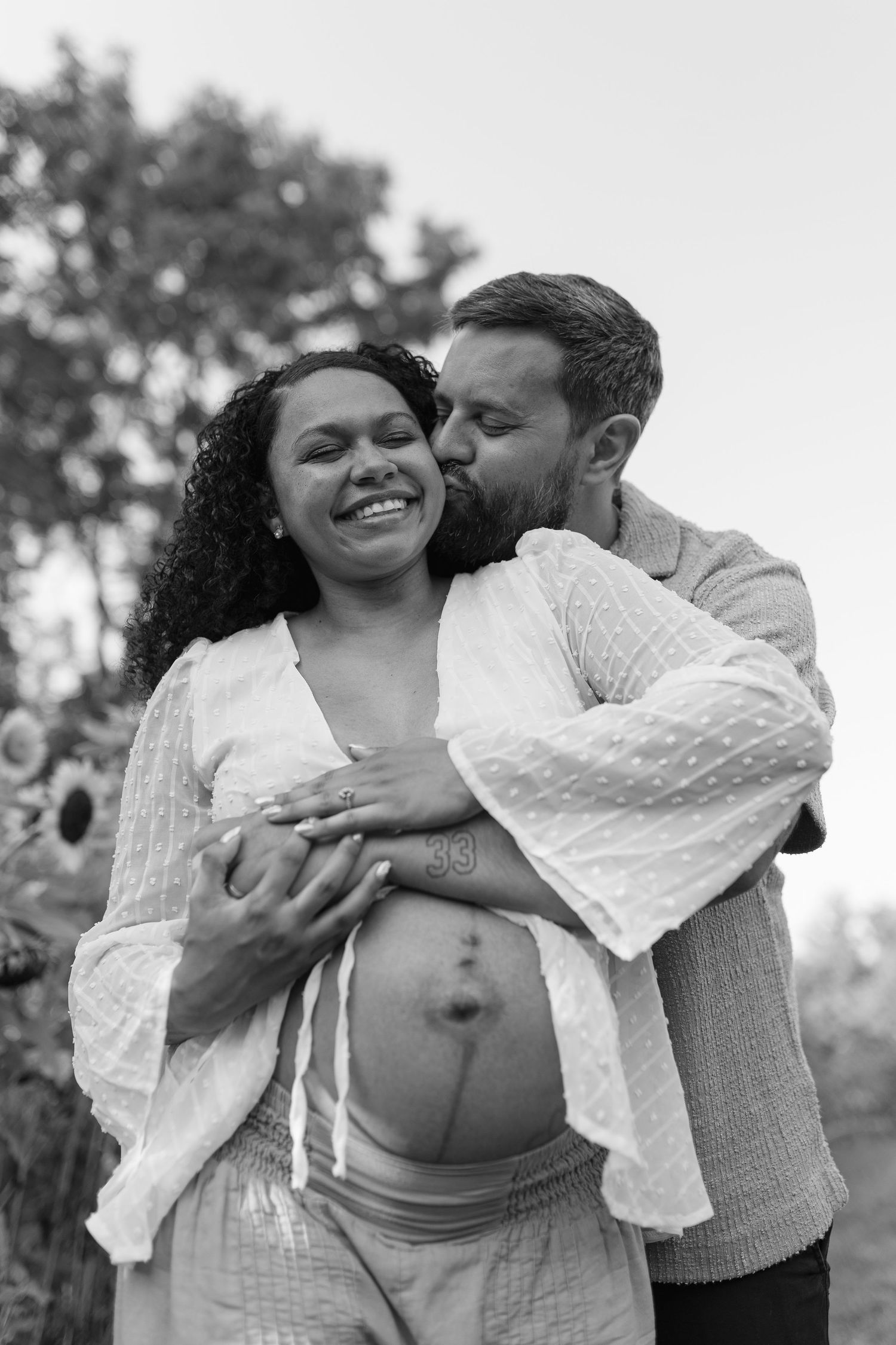Black and white maternity photo of a smiling pregnant woman being kissed on the cheek by her partner outdoors.