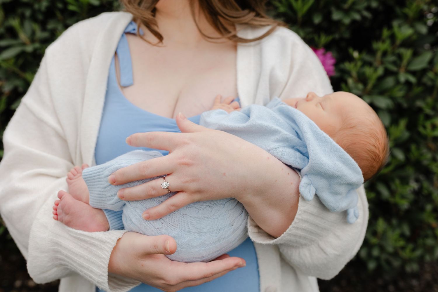 Mother in blue dress and white cardigan cradling sleeping newborn baby outdoors.