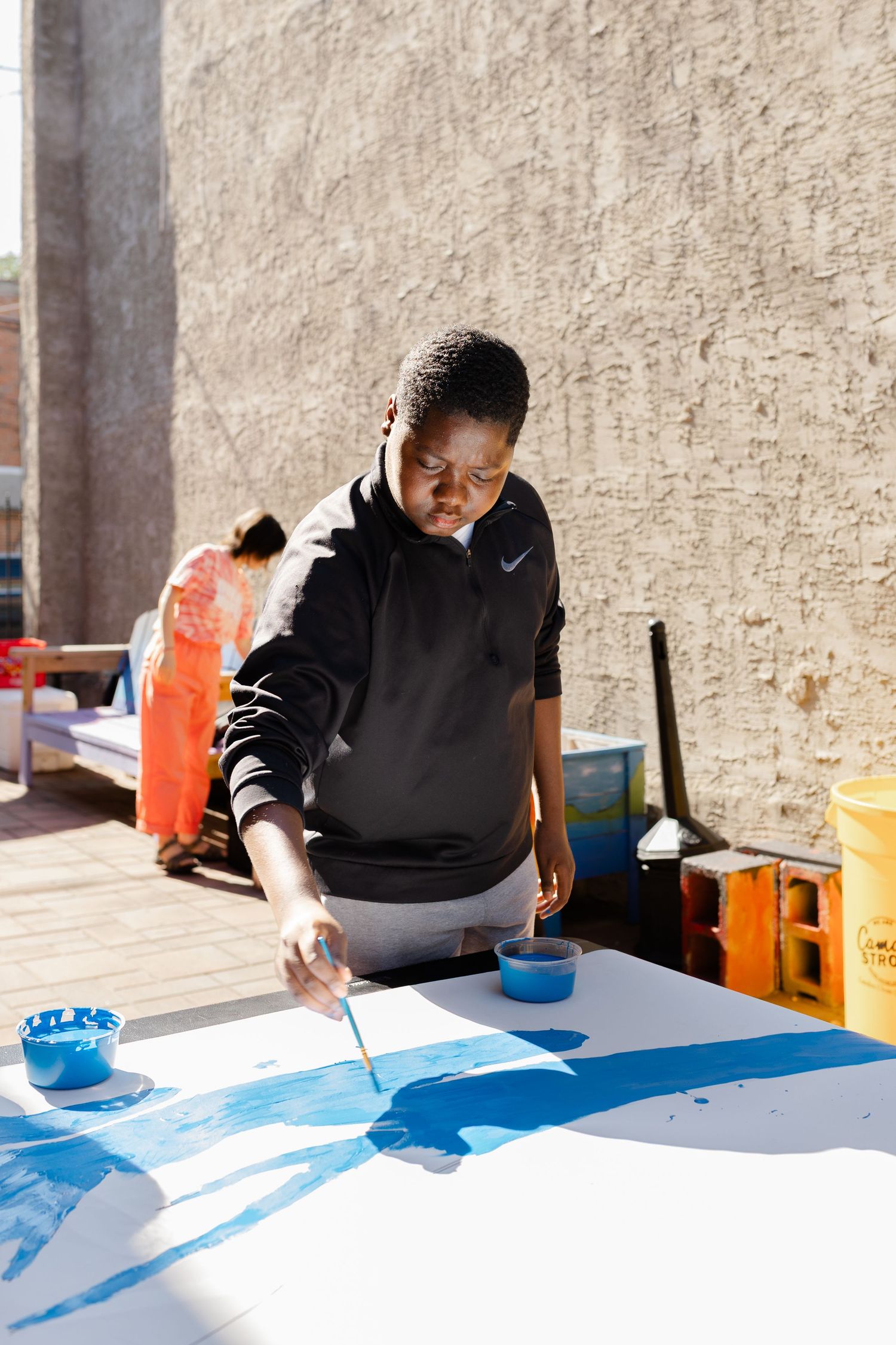 Young artist in black Nike hoodie painting large blue abstract artwork on white canvas outdoors.
