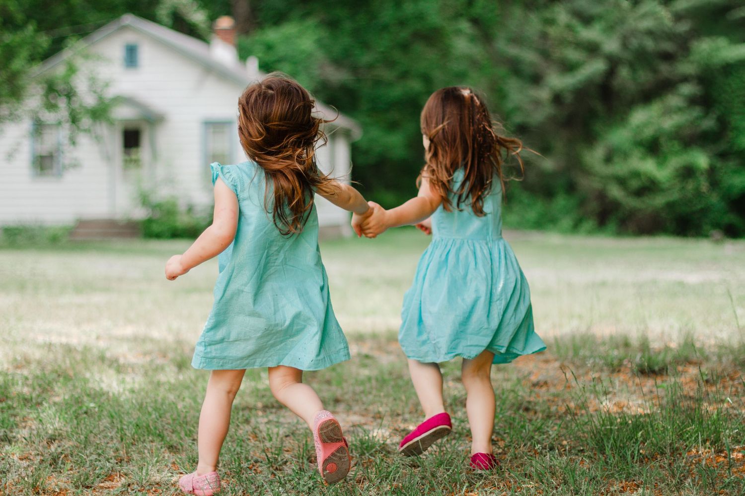 Two young girls in matching teal dresses hold hands while running through a grassy yard.