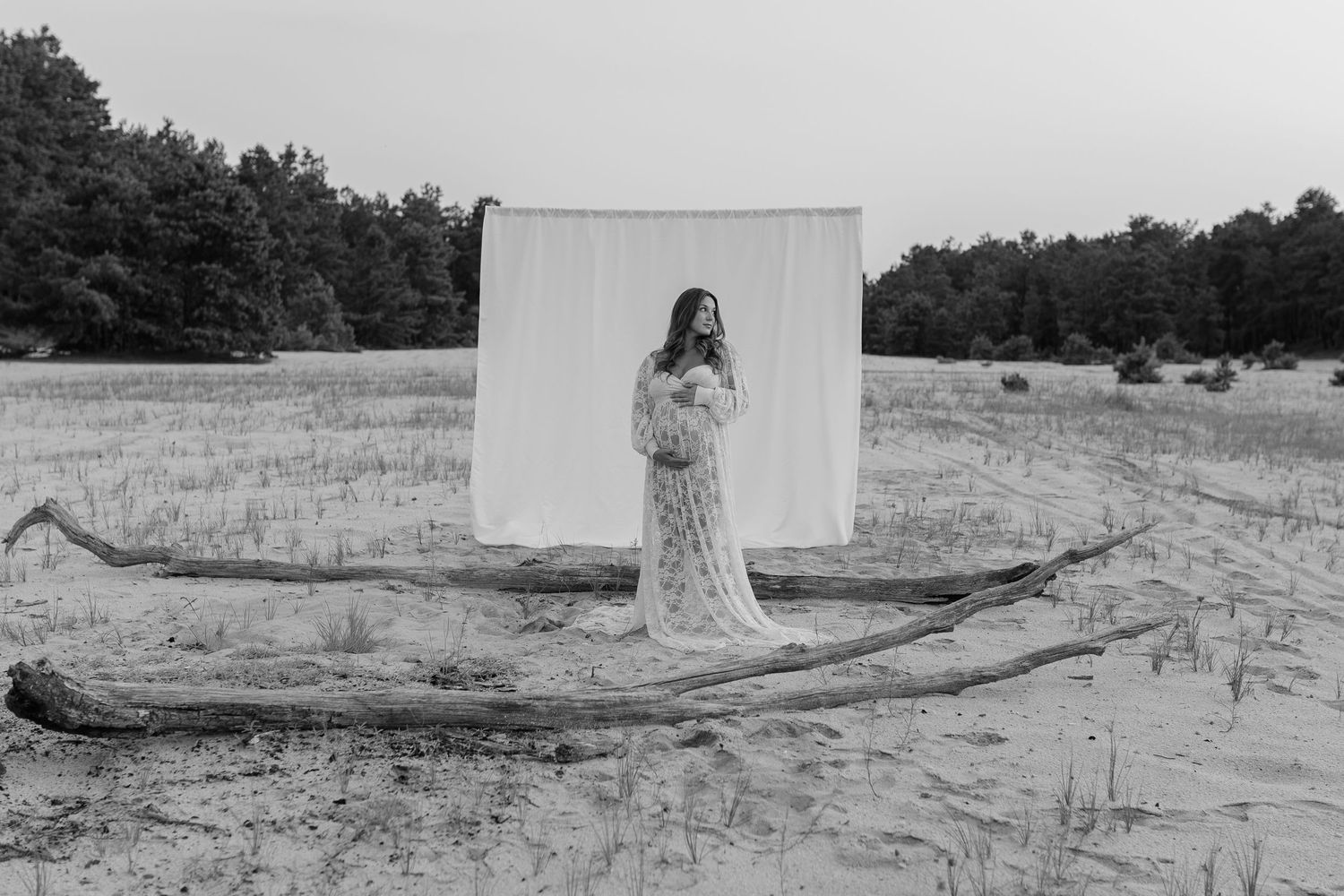 Pregnant woman in white lace gown stands before white backdrop on sandy field in black and white photo.