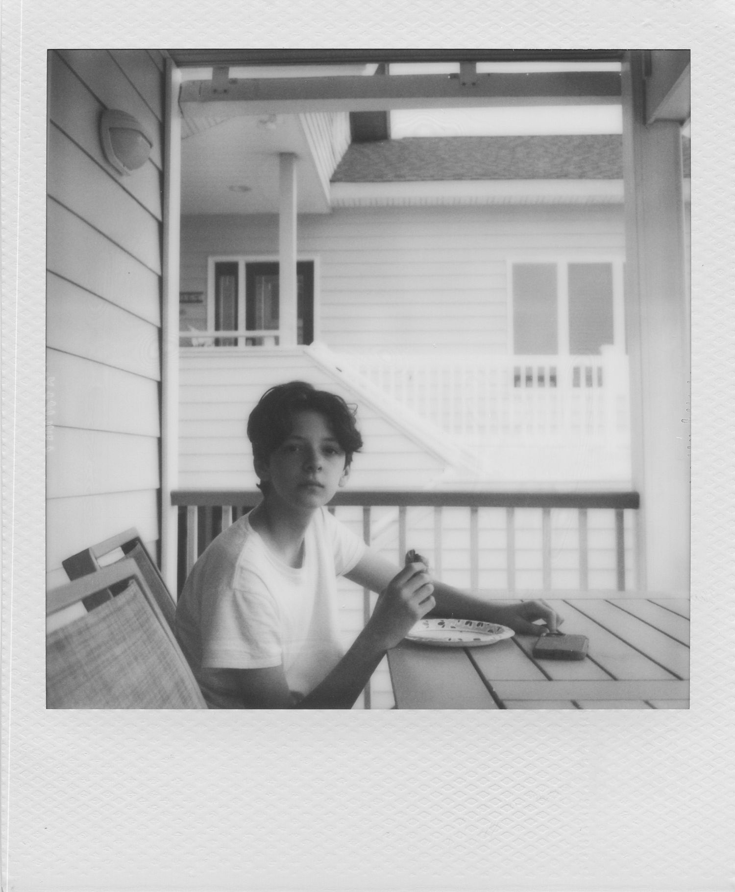 Black and white Polaroid photo of a young person sitting at an outdoor table on a porch, holding a cup.