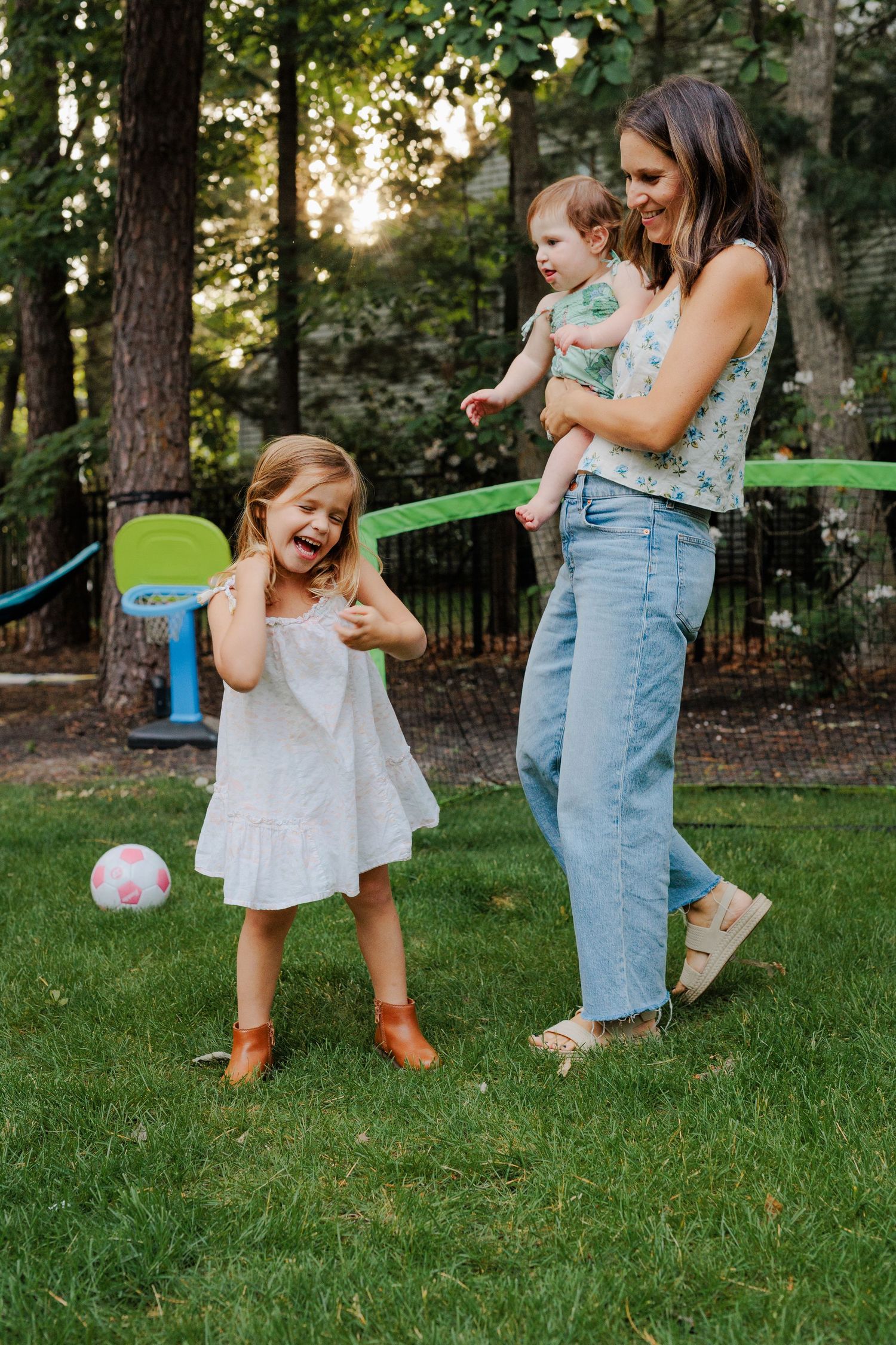 Smiling mother holding baby while young daughter laughs and kicks a pink ball in backyard.