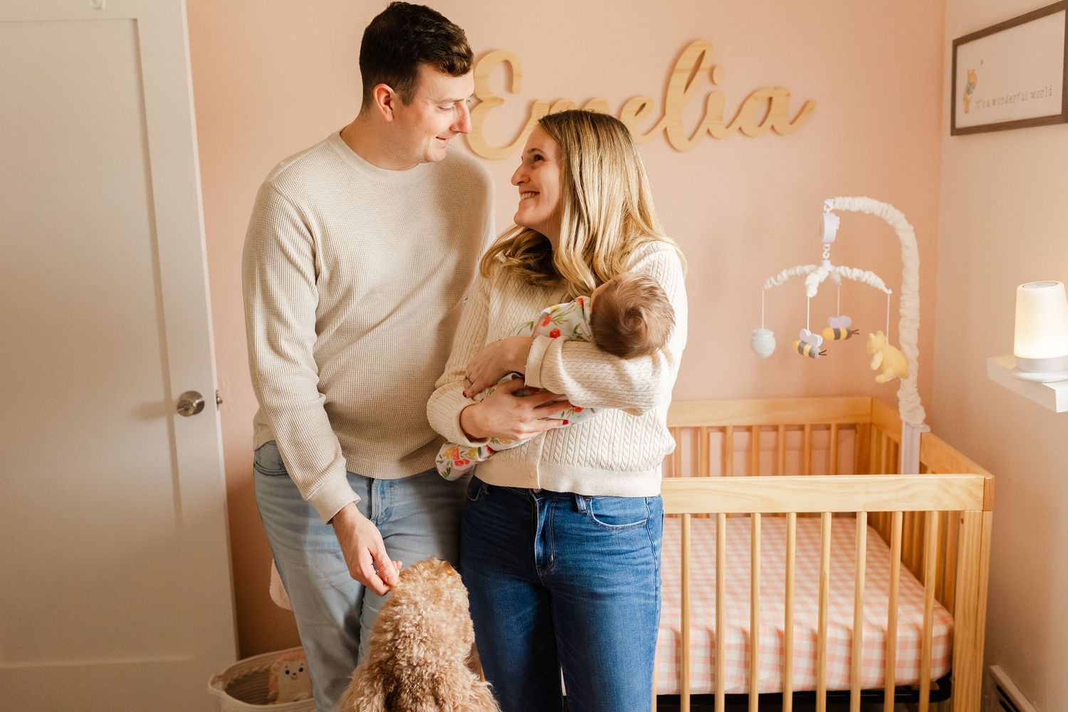 Happy couple in nursery with pink walls holds newborn baby near wooden crib with name decor.