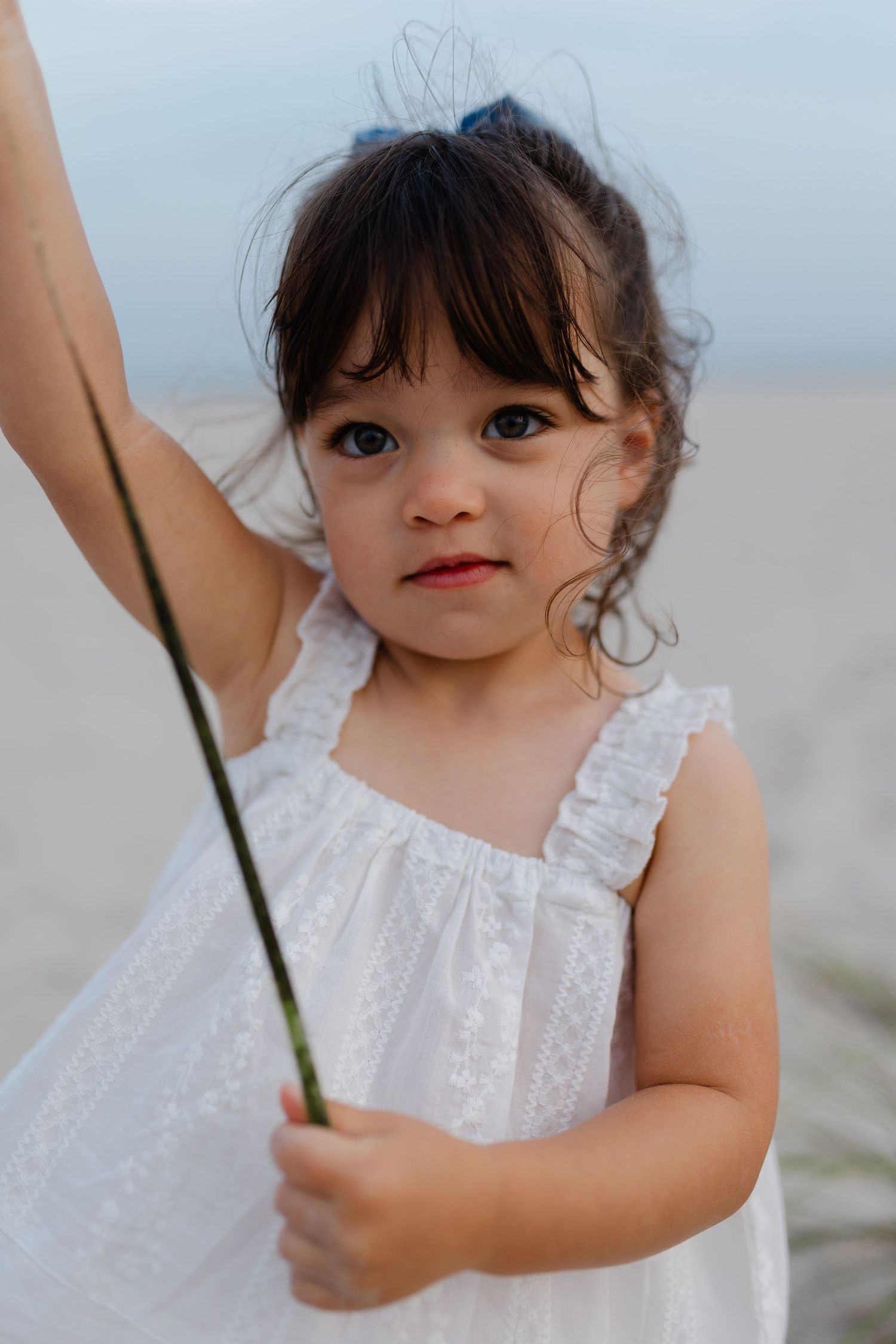 A young girl in a white lace dress holds a green stem at the beach, looking curiously at the camera.