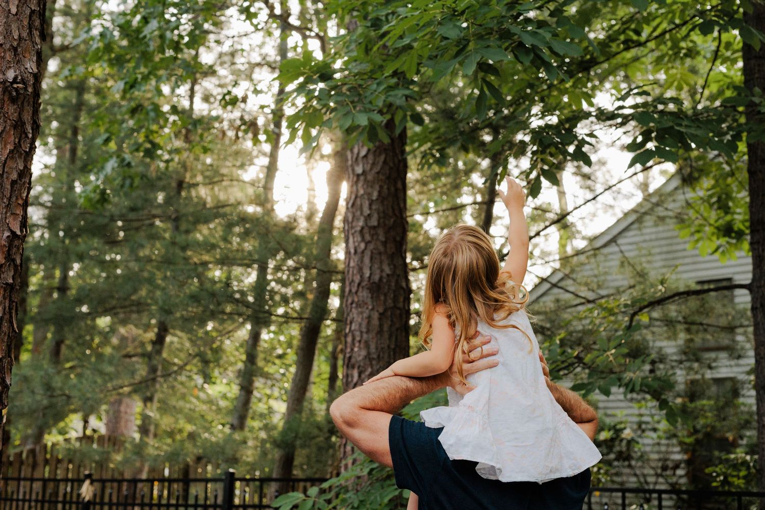 Child reaching up toward trees while being carried on an adults back in a lush green backyard.