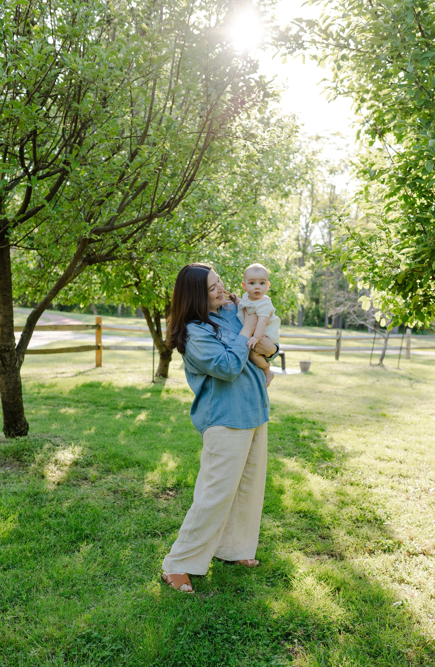 Mother lifting and gazing at her baby in a sunlit green park with trees in background.