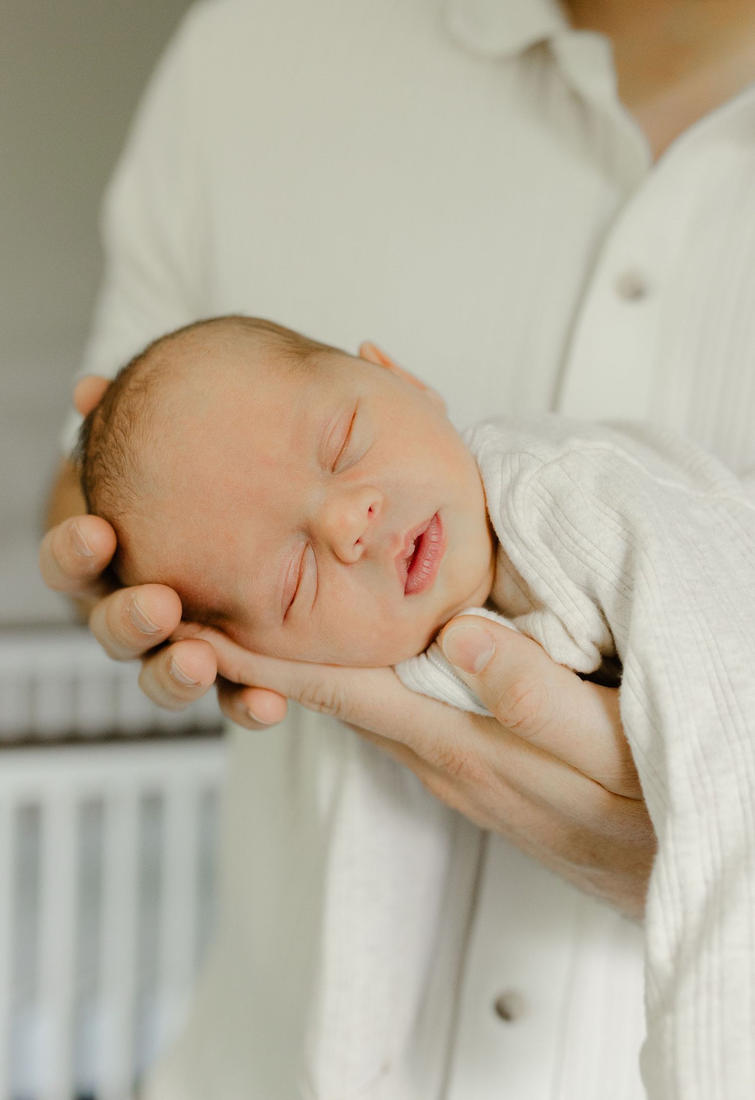 Peaceful sleeping newborn baby in white swaddle cradled gently in fathers hands during photoshoot.