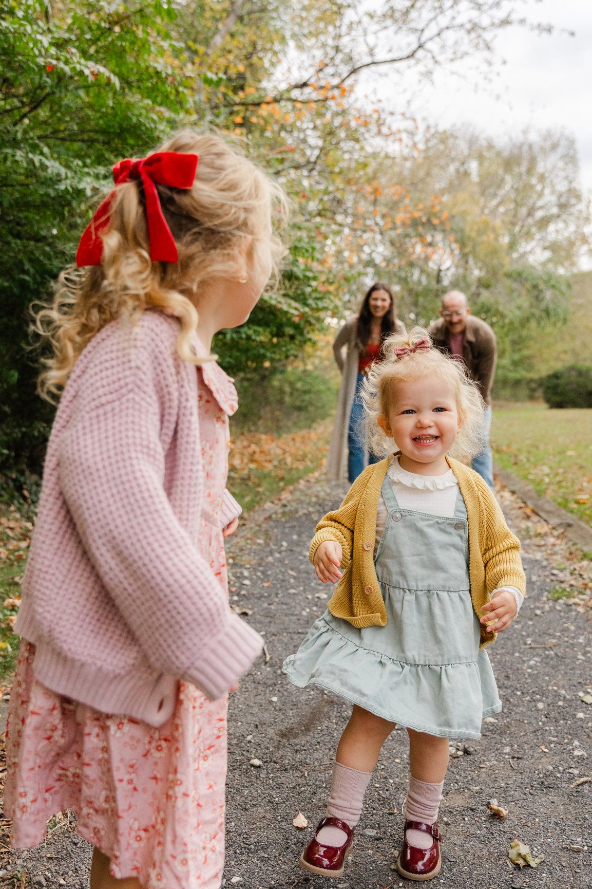 Two young sisters laughing on a path in autumn park with parents smiling in background.