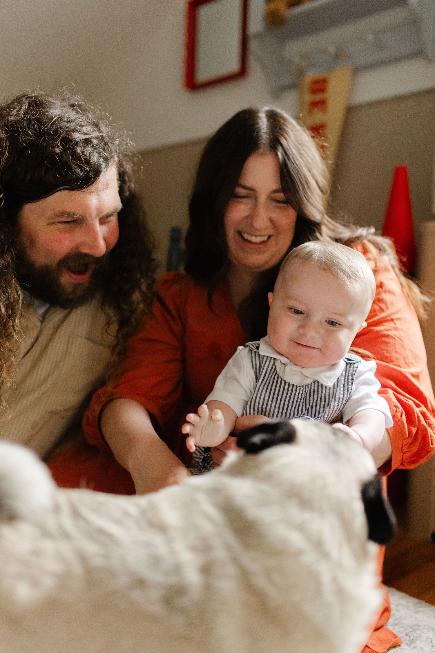 Laughing parents watching their baby reach toward their pug dog in a cozy home setting.