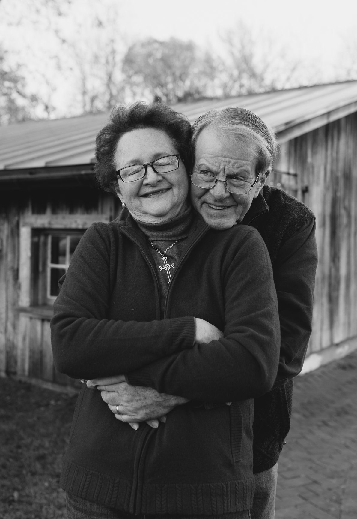 Black and white portrait of a smiling elderly couple embracing warmly outside a rustic wooden building.