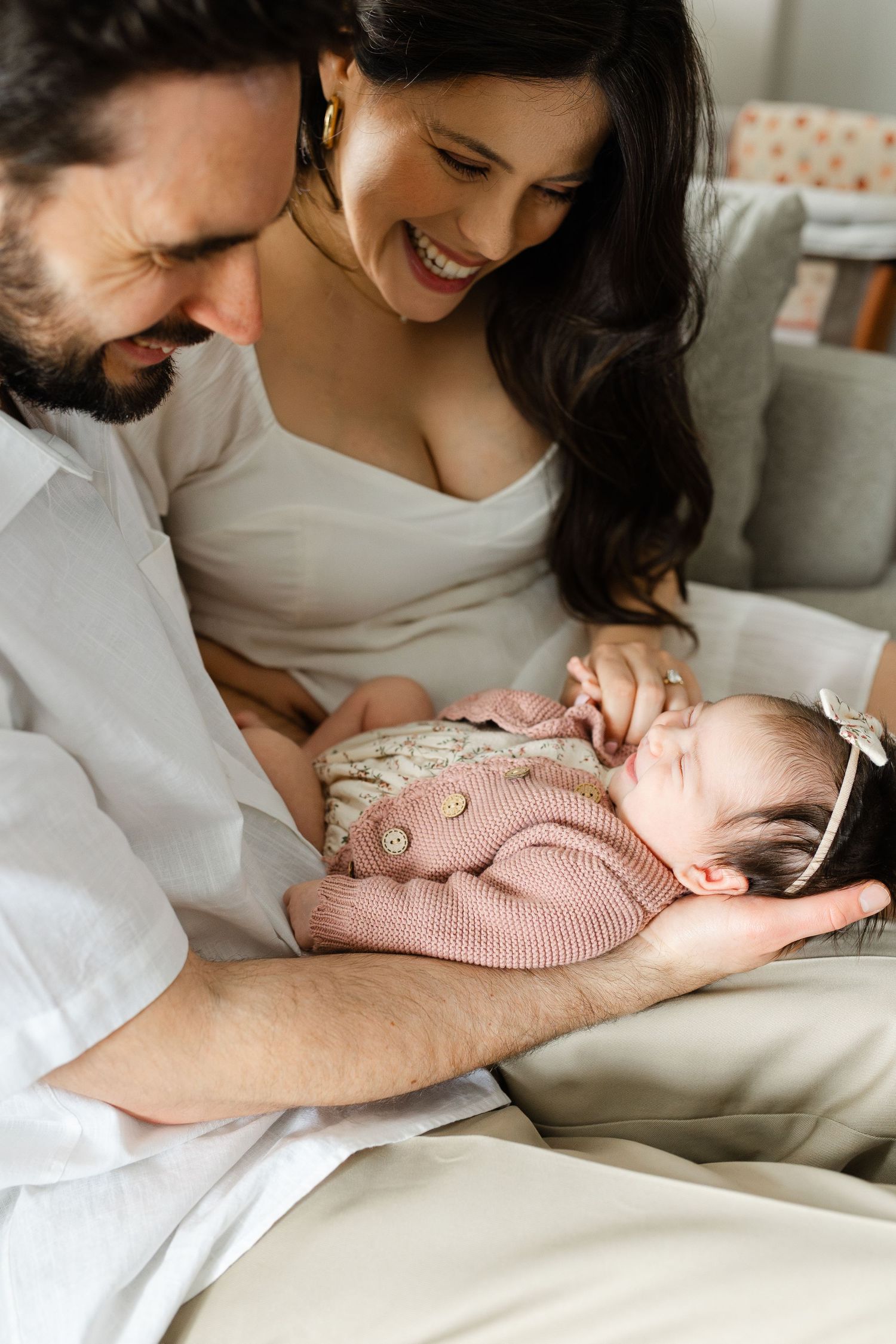 Smiling parents in white clothing lovingly gazing at their sleeping newborn baby in pink outfit.
