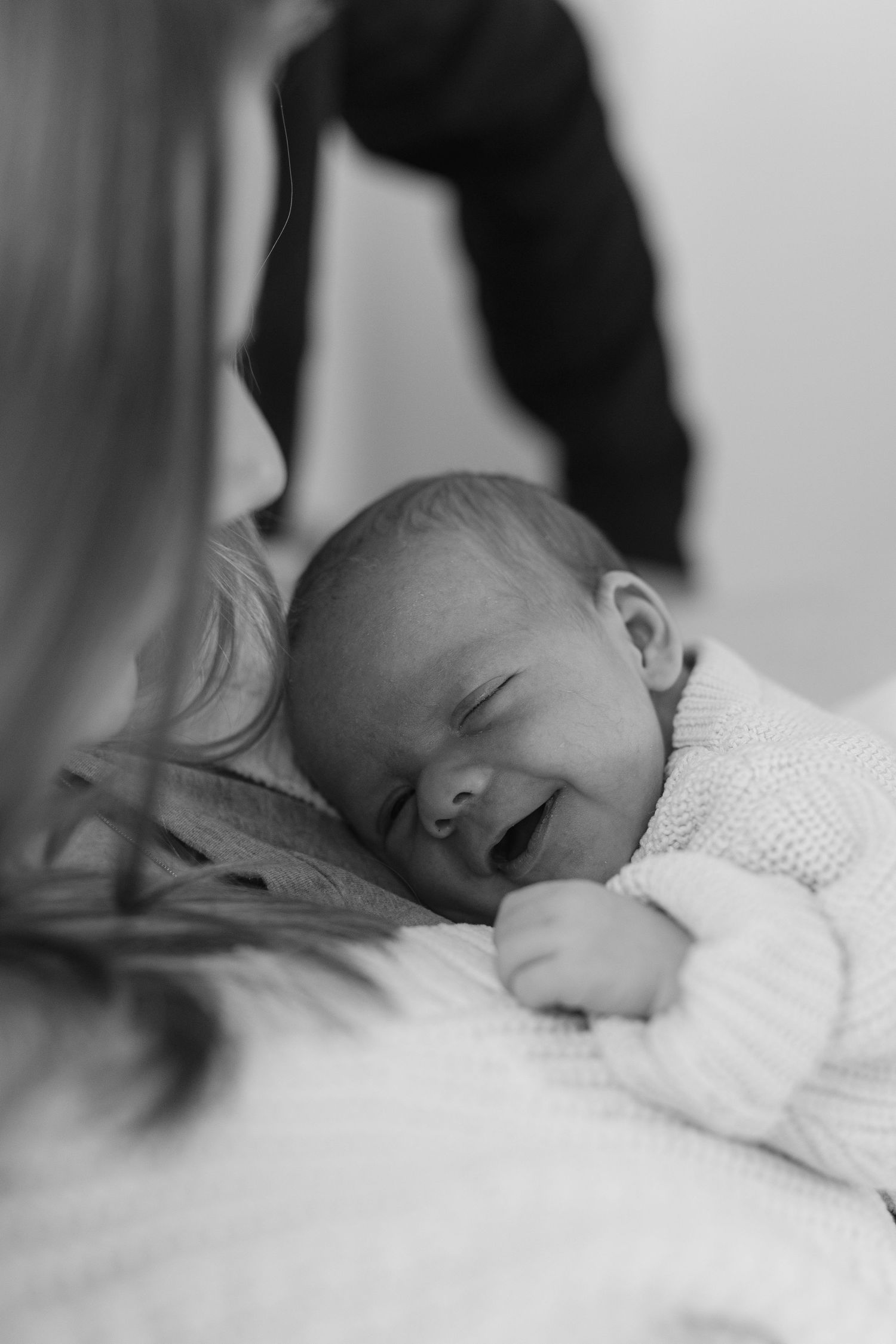 Black and white photo of smiling newborn baby lying on soft blanket while mother looks on closely.