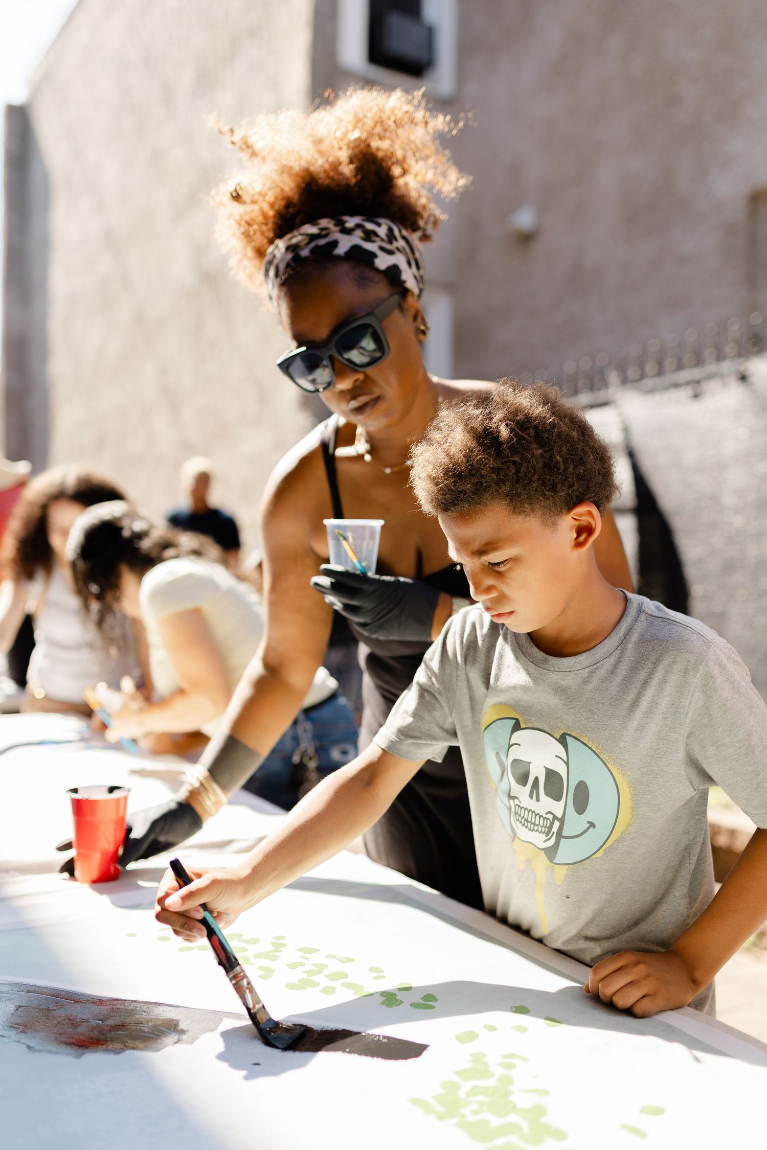 A woman and boy painting together on a large canvas outdoors at a community art event.